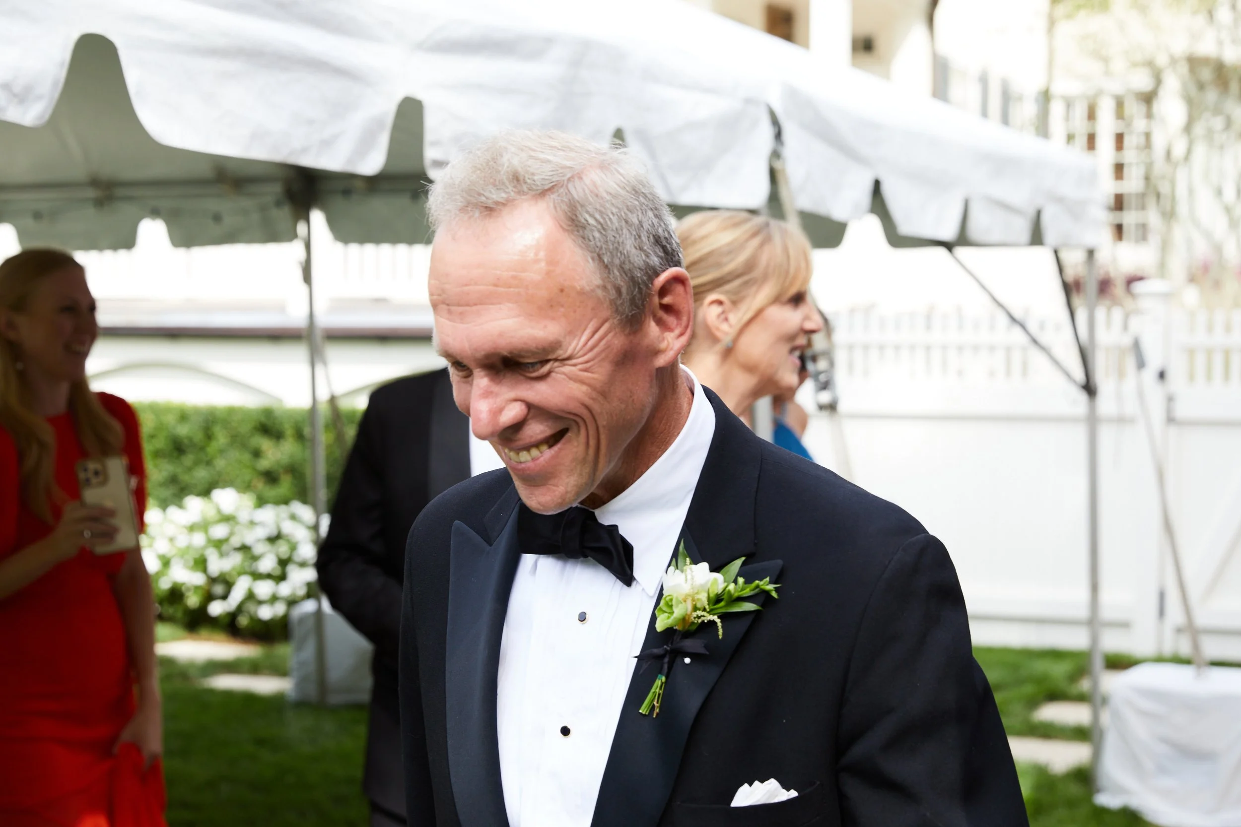 A smiling man in a black tuxedo with a white shirt and black bow tie, wearing a boutonnière, at an outdoor event, with other people and a white tent in the background.