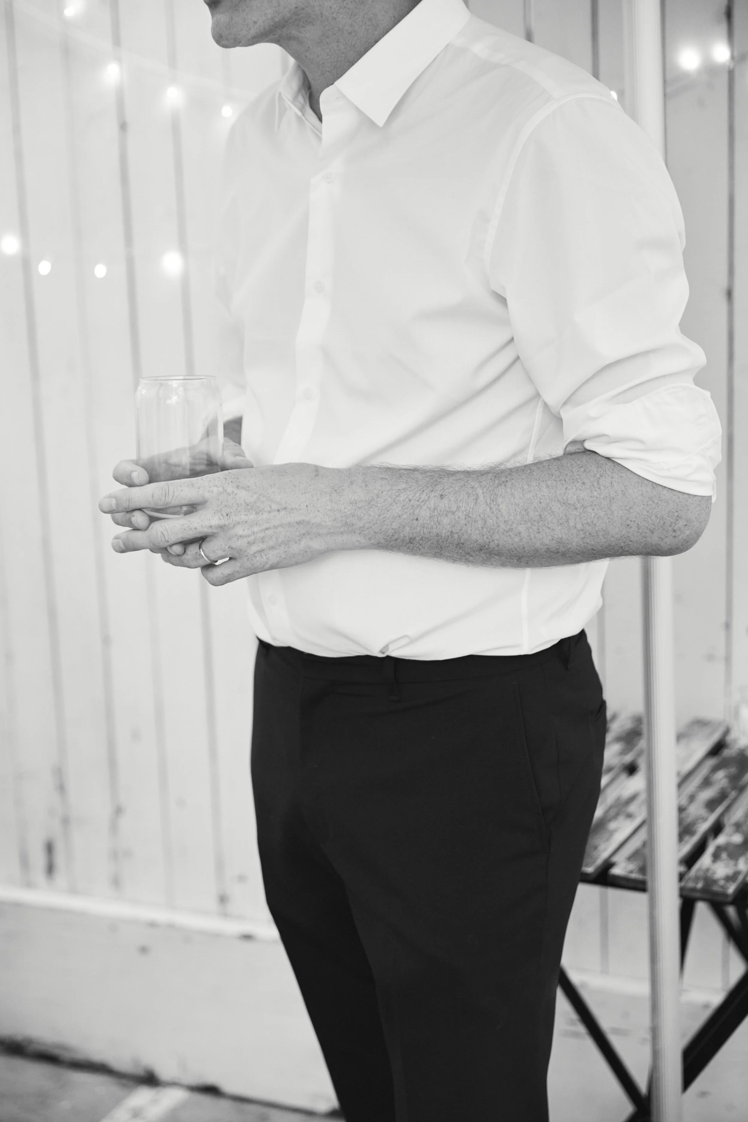 A man dressed in a white shirt and black pants holding a glass of water, standing in front of a wooden panel wall.