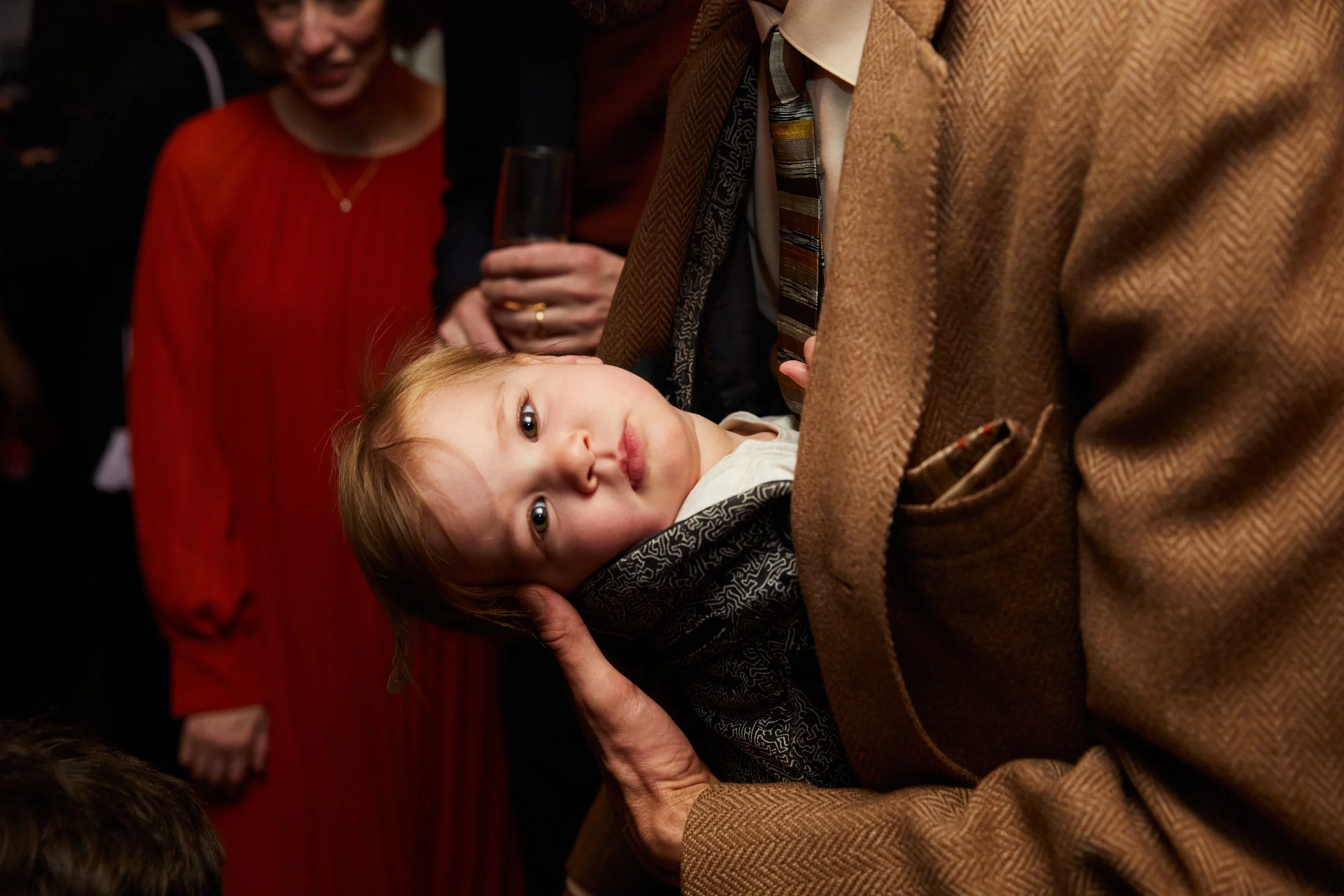 A young boy with light brown hair lying on an adult's arm, looking directly at the camera. The adult is wearing a brown jacket and patterned tie. In the background, there is a woman in a red dress and other people at a social gathering.