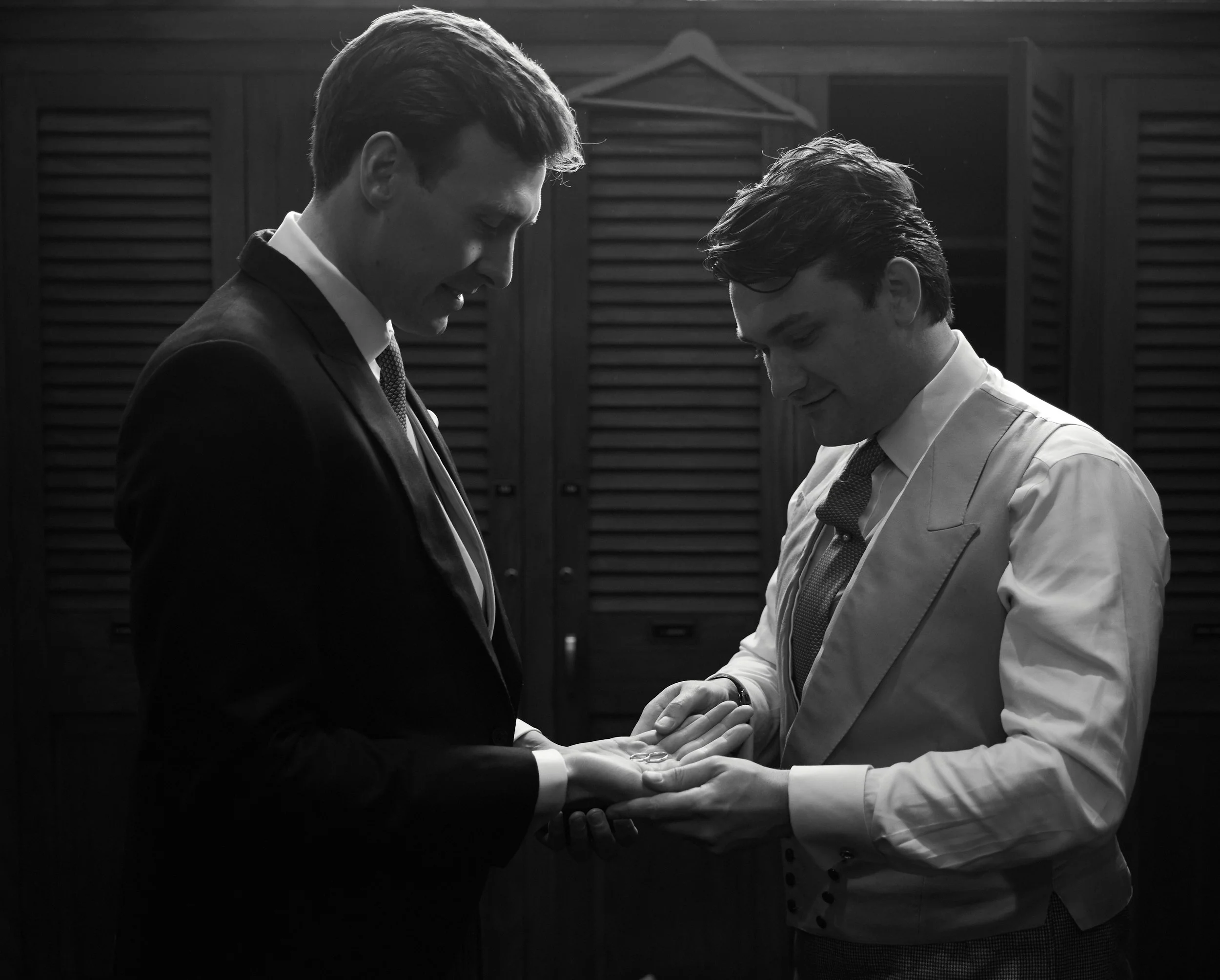 Two men in suits, one in a dark suit and one in a light suit, standing close together and exchanging rings in a solemn moment.
