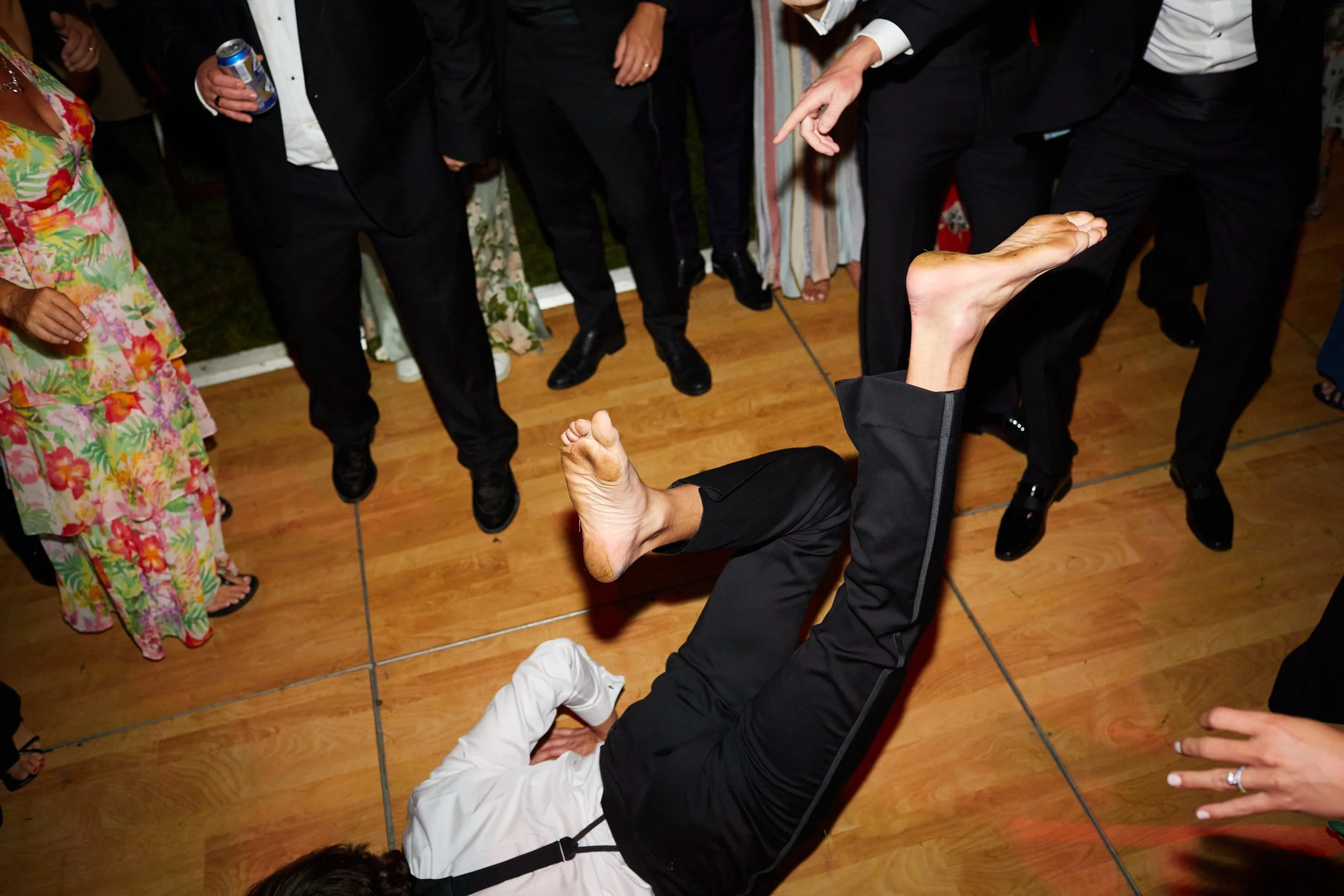 Person lying on the dance floor with legs up, surrounded by standing guests at a celebration or party.