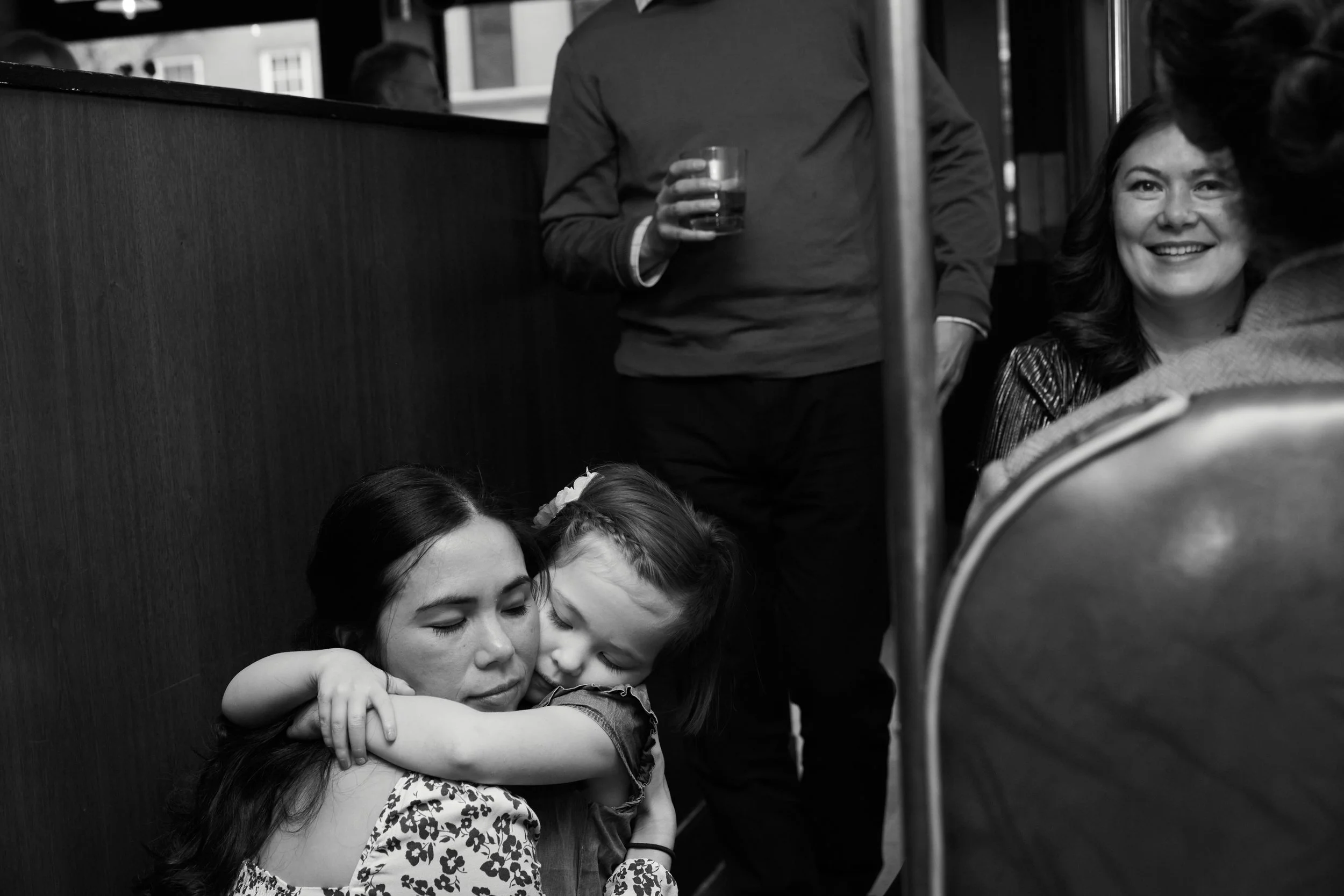 A young girl hugging a woman with closed eyes, both with dark hair, in a social setting with people standing around.