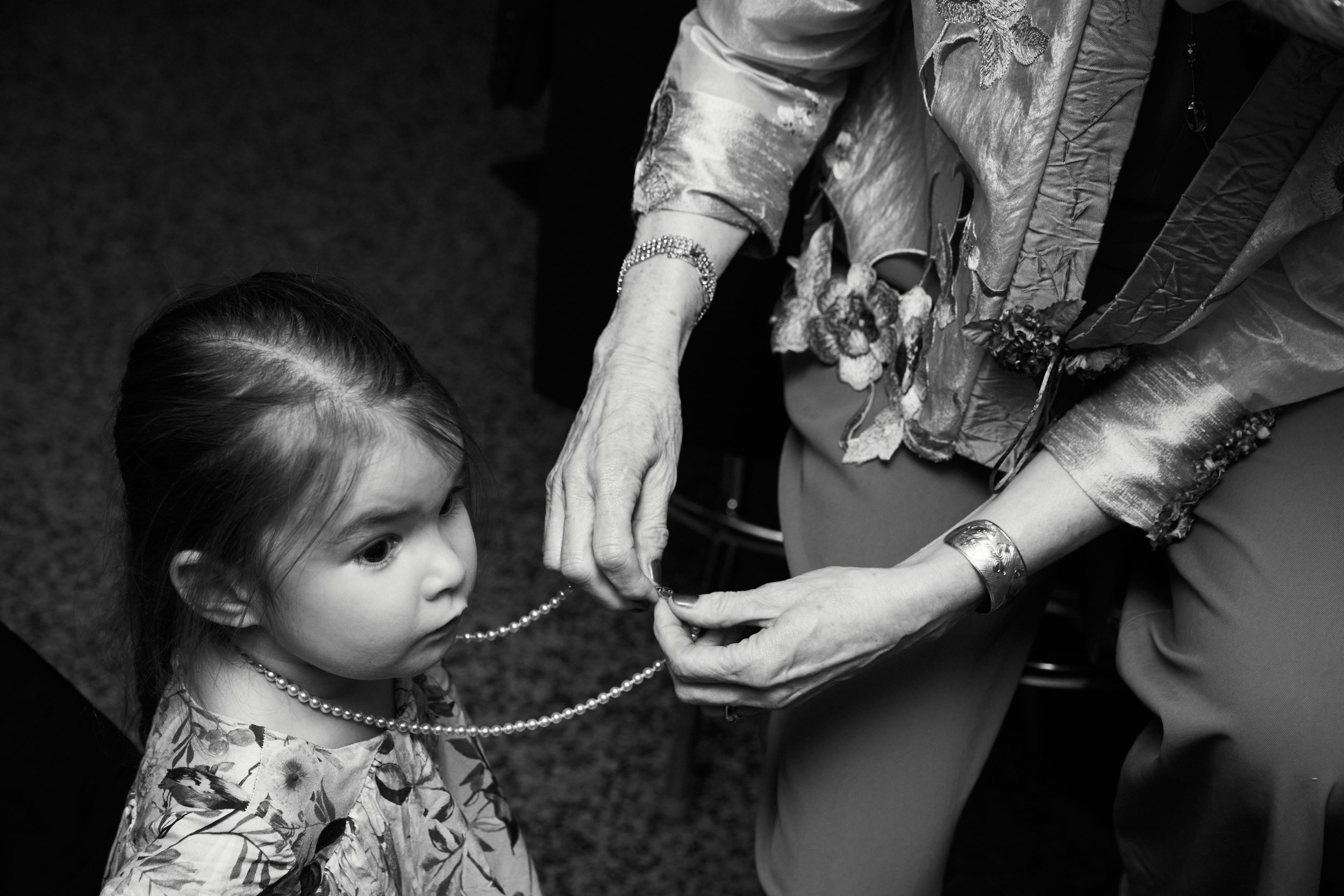 An elderly woman is fastening a pearl necklace around a young girl's neck. The elderly woman is wearing a floral dress with embroidered details and bracelets. The girl has dark hair tied back and is wearing a patterned top, looking attentively at the
