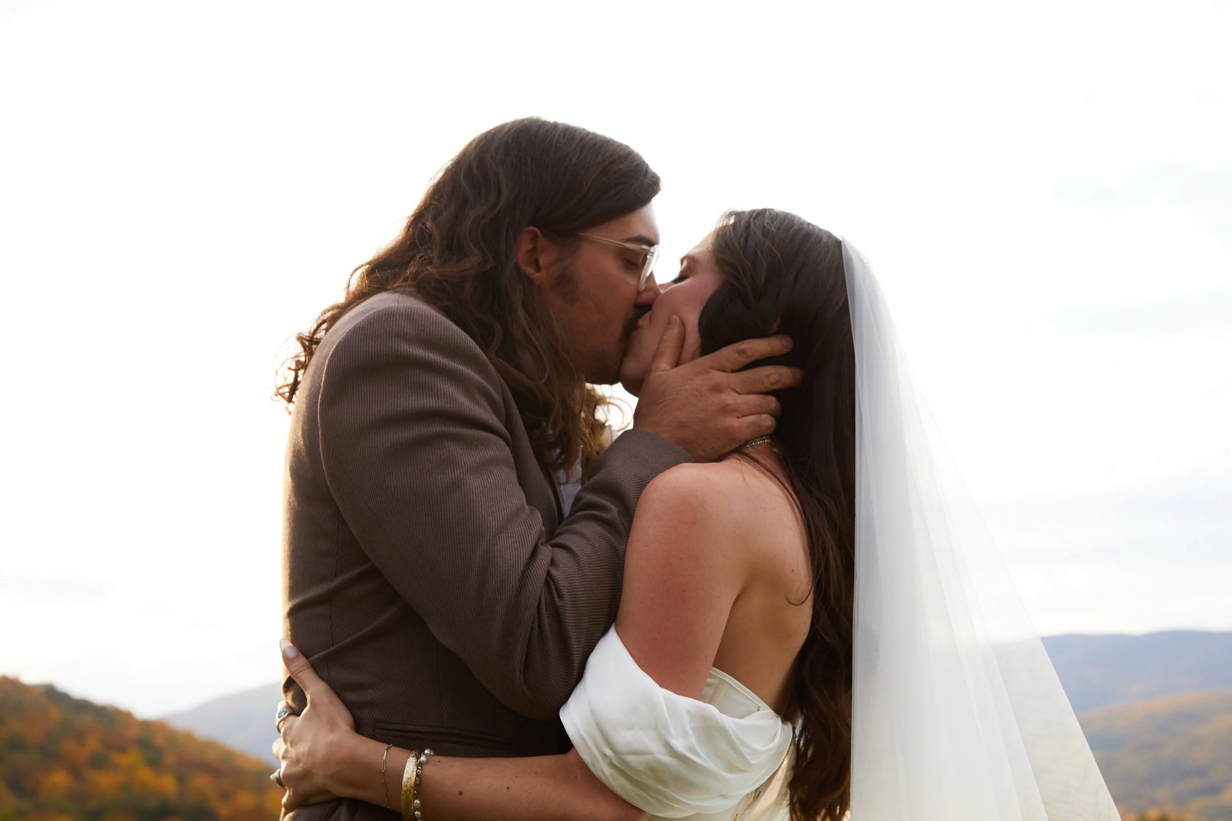 A couple in wedding attire sharing a kiss outdoors, with mountains and sky in the background.