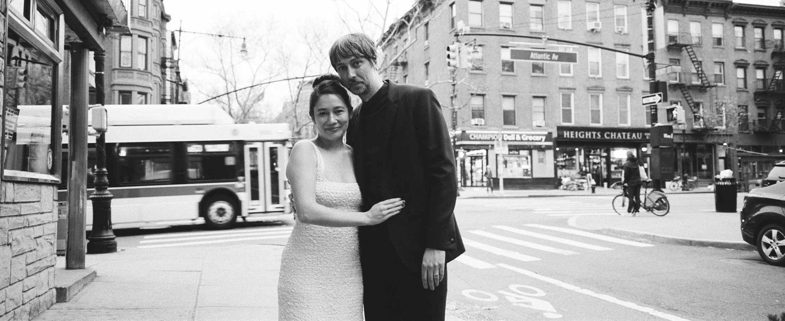 A black-and-white photo of a couple standing on a city sidewalk with a street, bus, and buildings in the background.