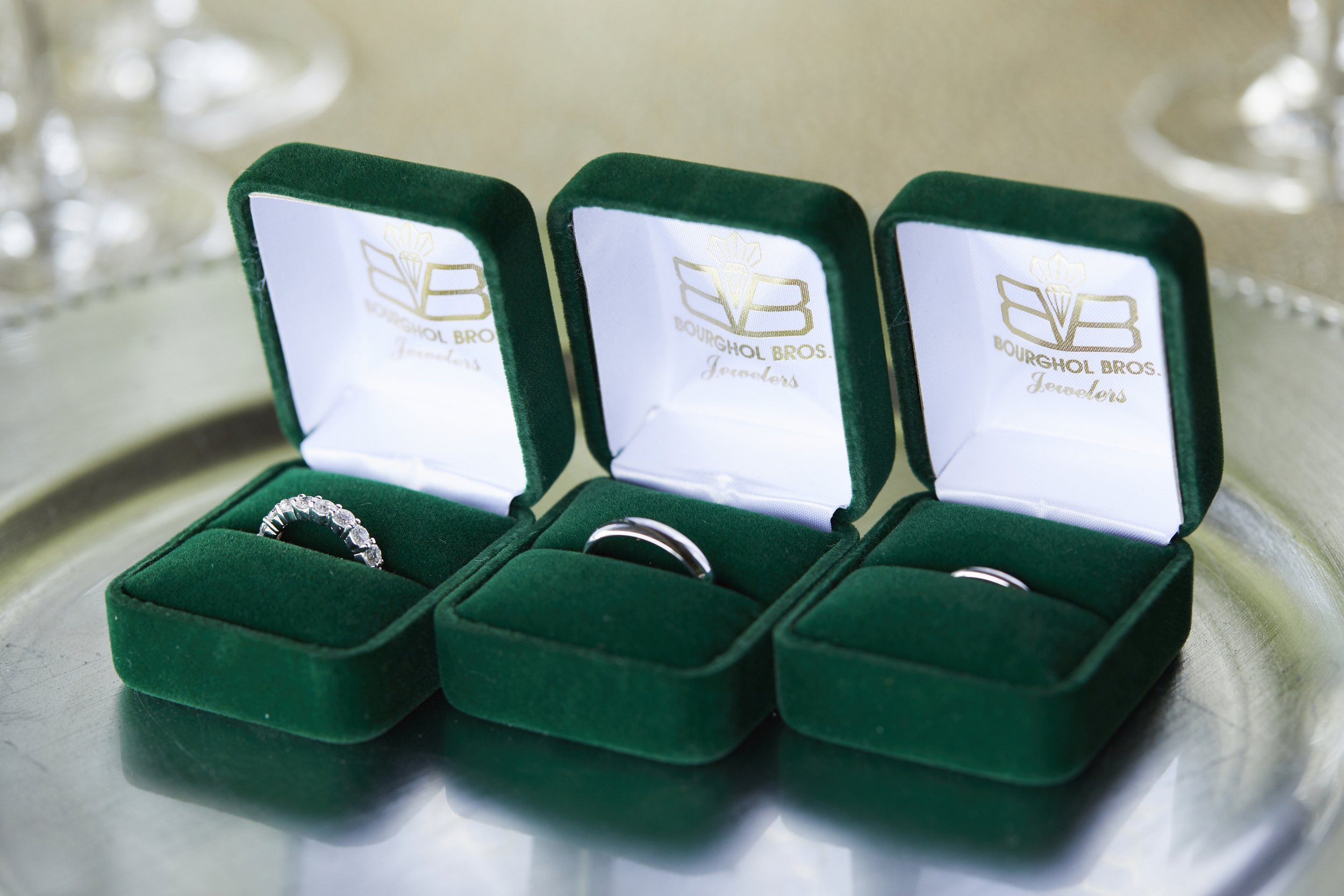 Three green jewelry boxes with rings inside, placed on a reflective surface. The boxes have white interior linings with the Bourghol Bros. Jewelers logo printed inside.