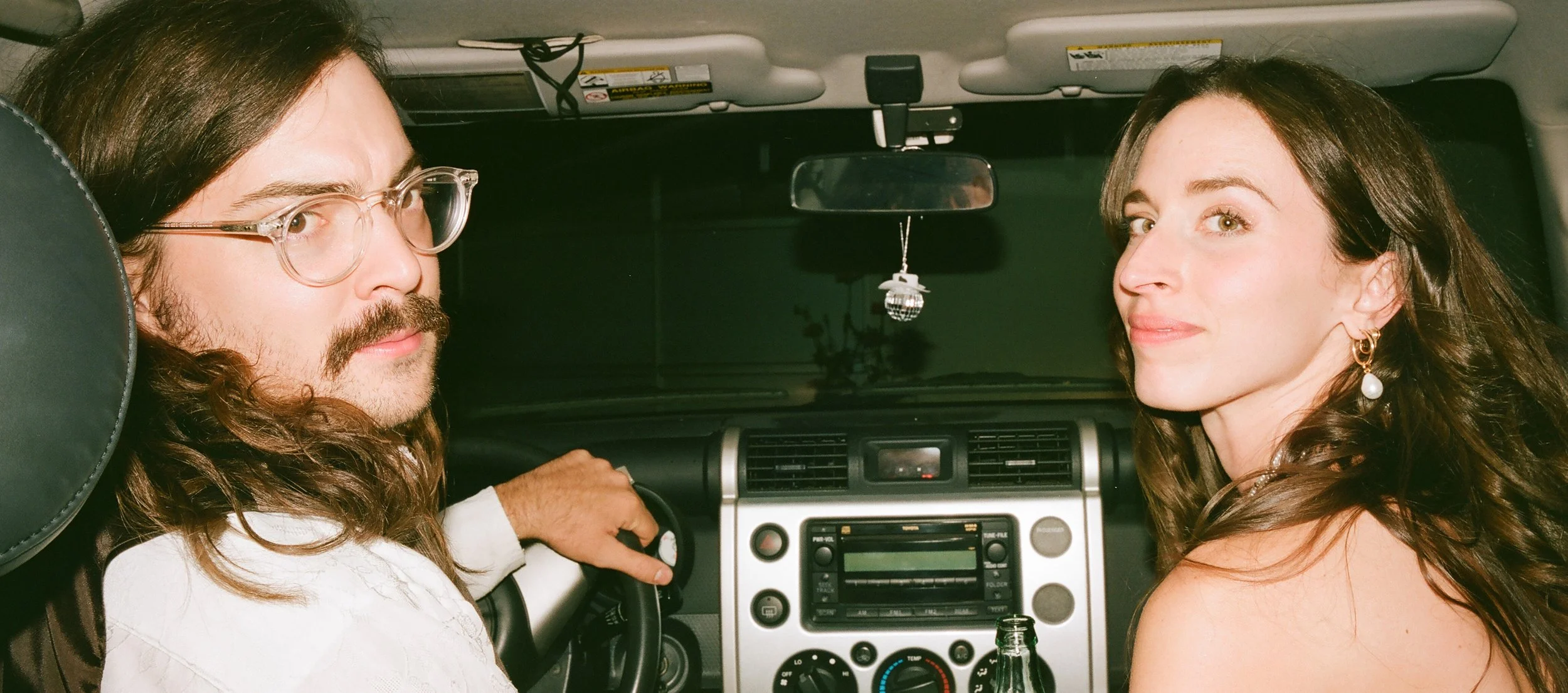 A man with long hair, glasses, and a beard driving a car, with a woman with long brown hair and earrings sitting in the passenger seat beside him.