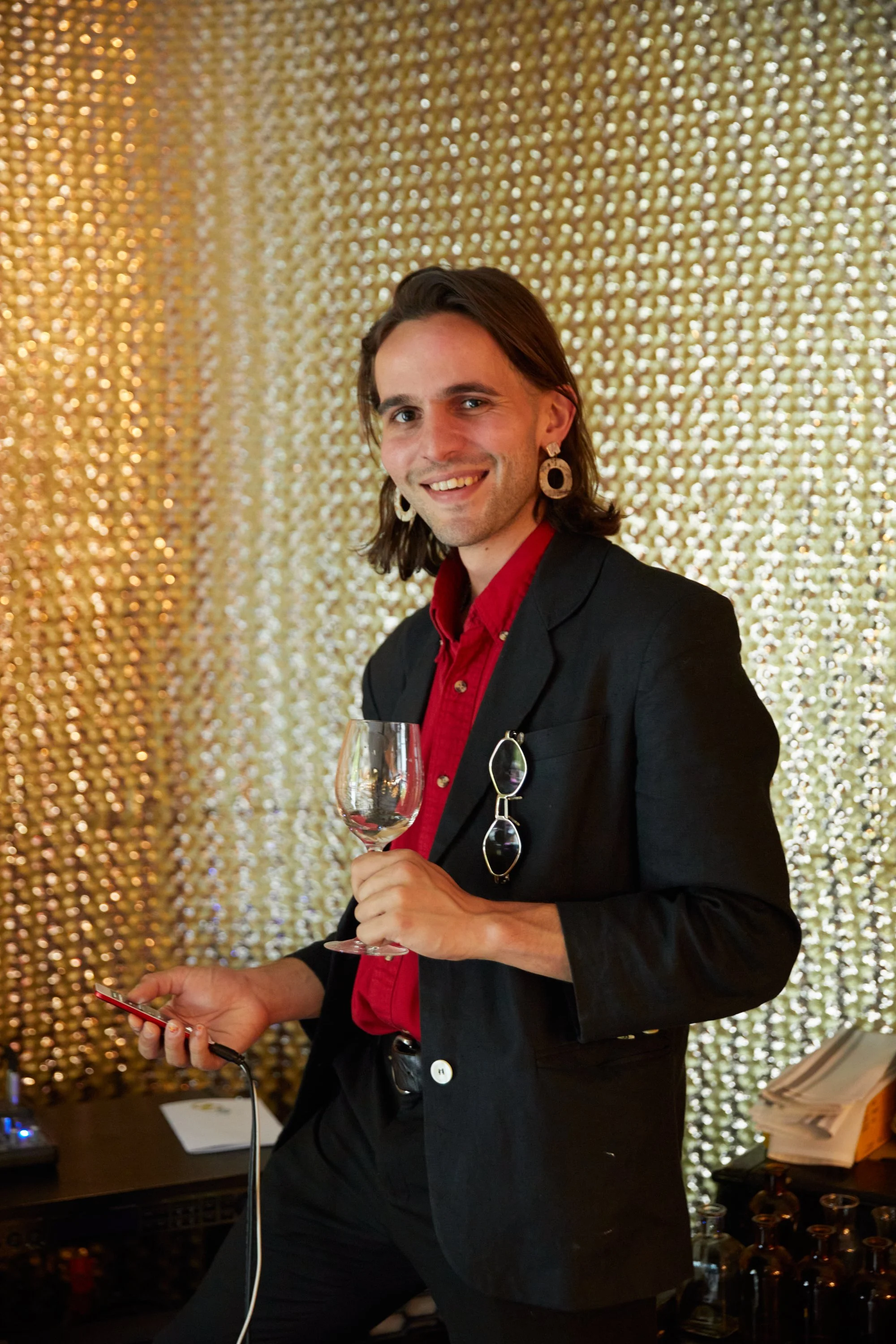 A man with shoulder-length brown hair, wearing a black blazer, red shirt, and large earrings, holding a wine glass and a phone in front of a gold sequin backdrop.