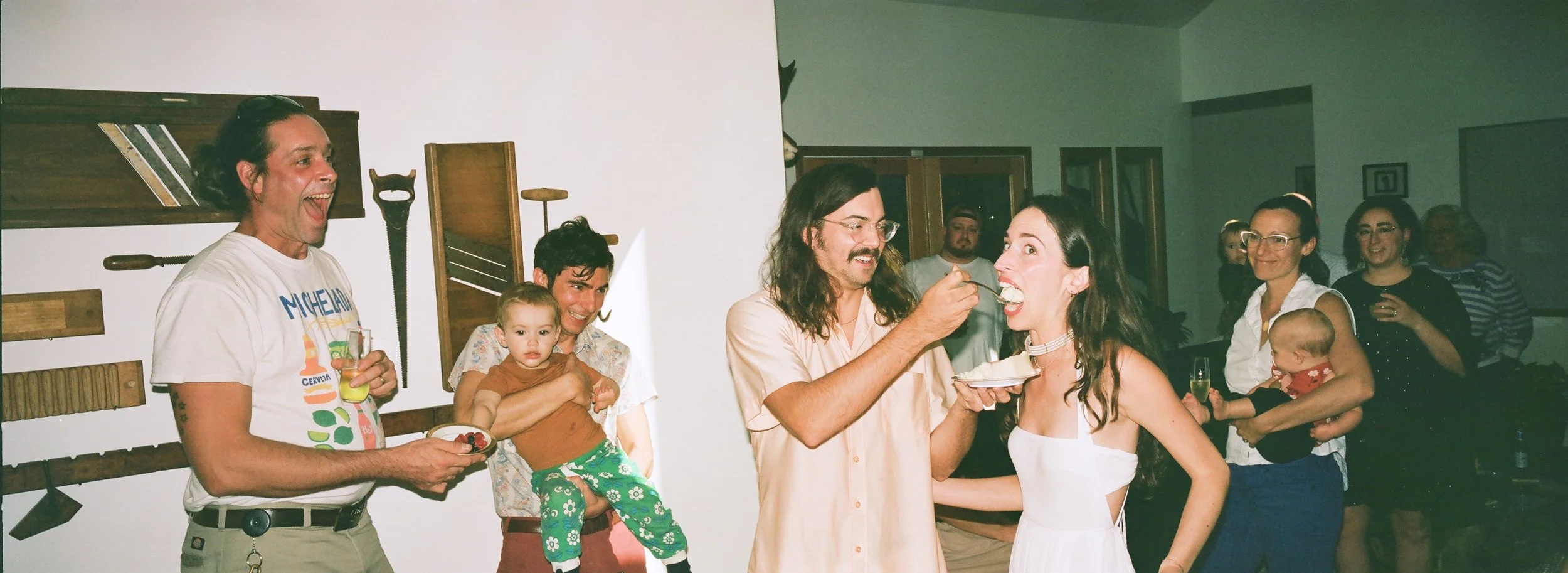 A group of people celebrating a birthday, with a woman wearing a white dress being fed cake, while others watch and smile. A man in a white shirt with colorful graphics and a man with long hair and glasses are among the guests.