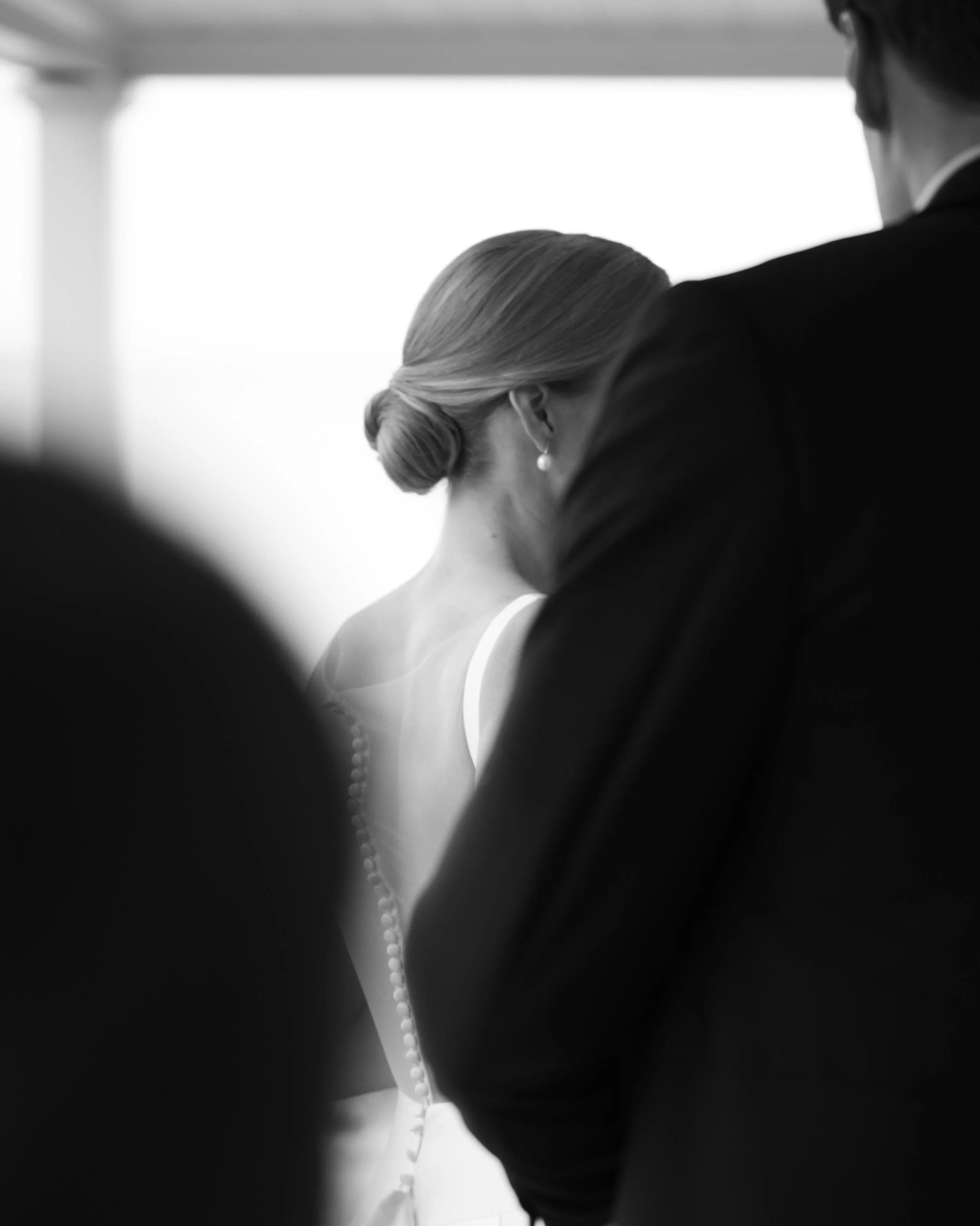 Black and white photo of a woman with her hair in a bun, wearing a dress with beads on the back, at a formal event, possibly a wedding, with two men in tuxedos nearby.