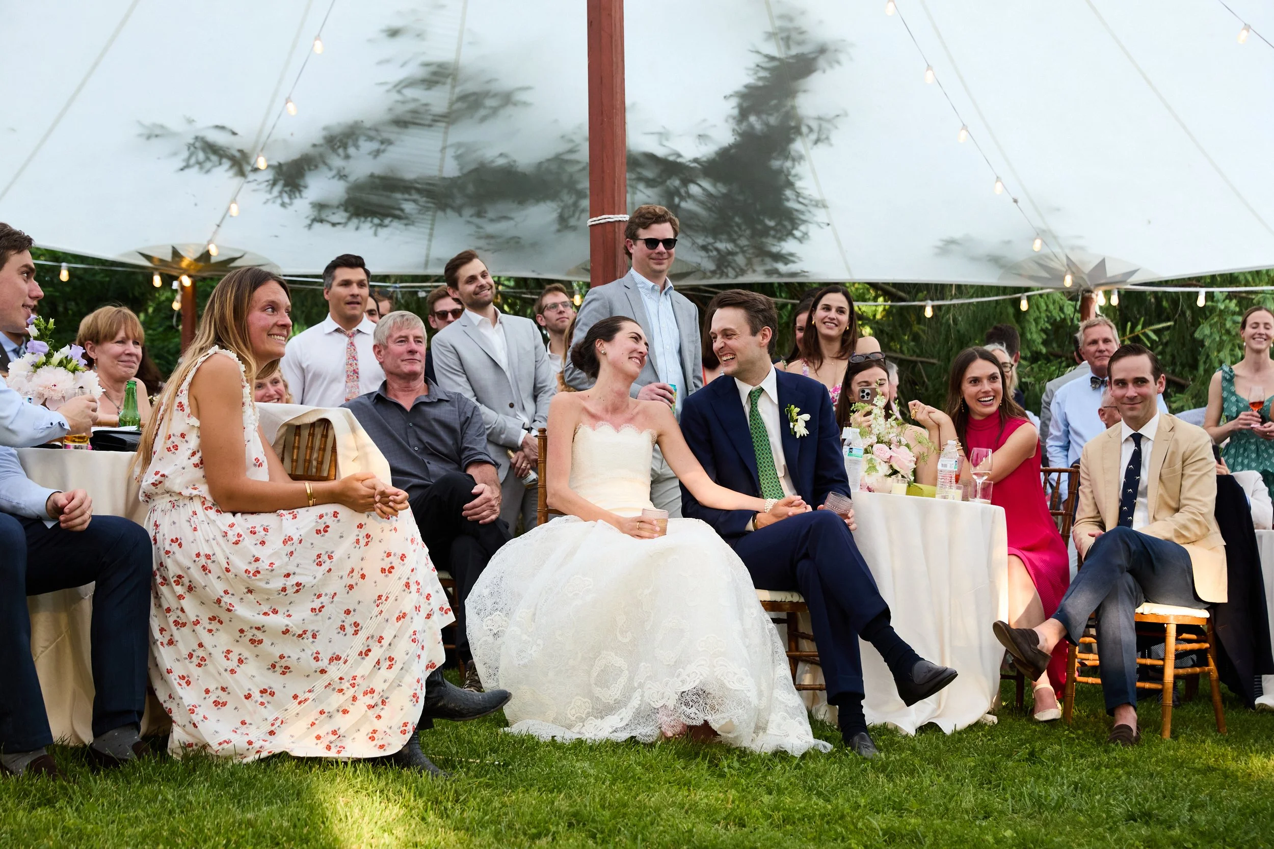 A bride and groom sitting at a table during a wedding reception, smiling and holding glasses, surrounded by friends and family in an outdoor setting with a canopy and string lights.