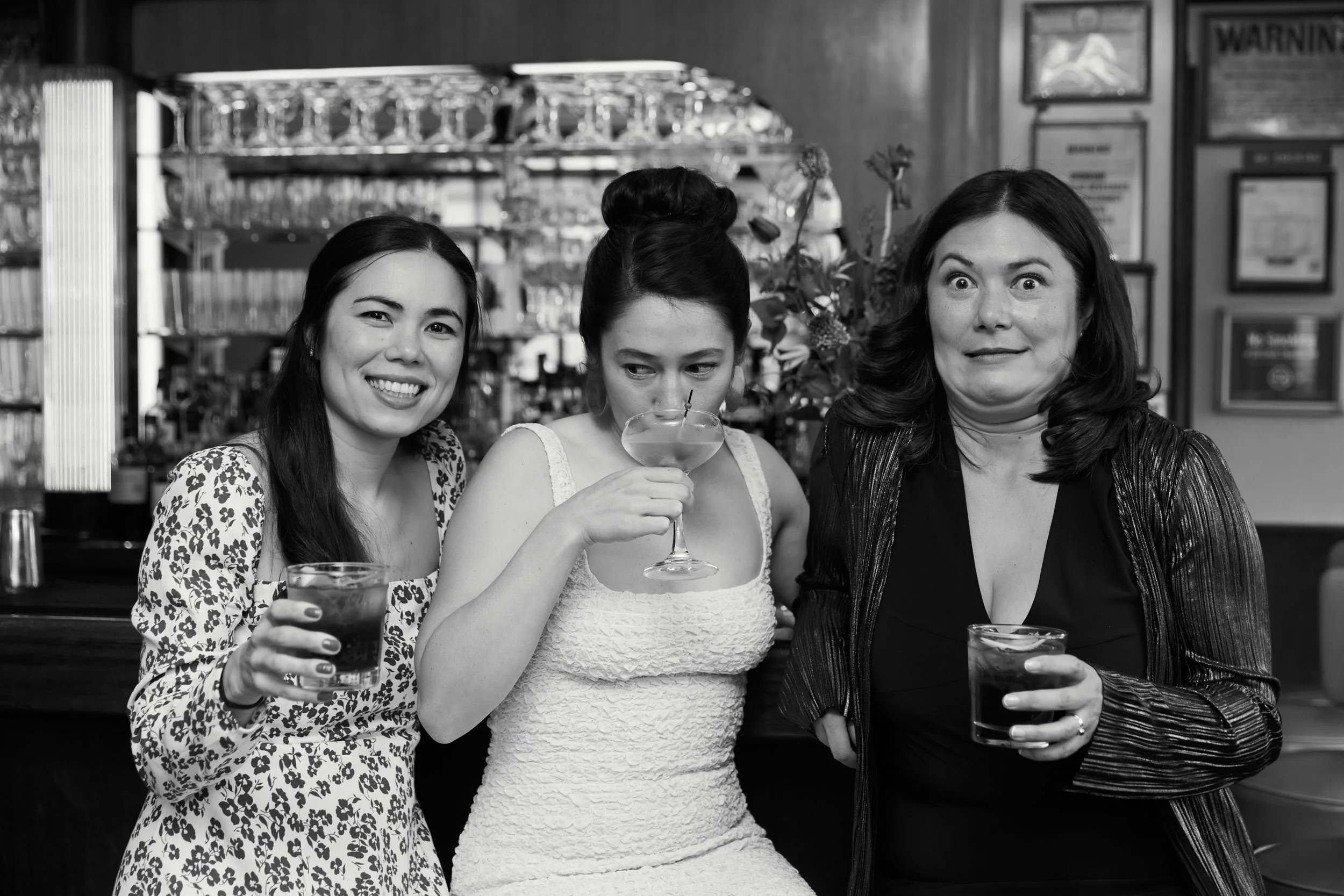Three women standing close together at a bar, holding drinks, with a background of glasses and framed posters on the wall.