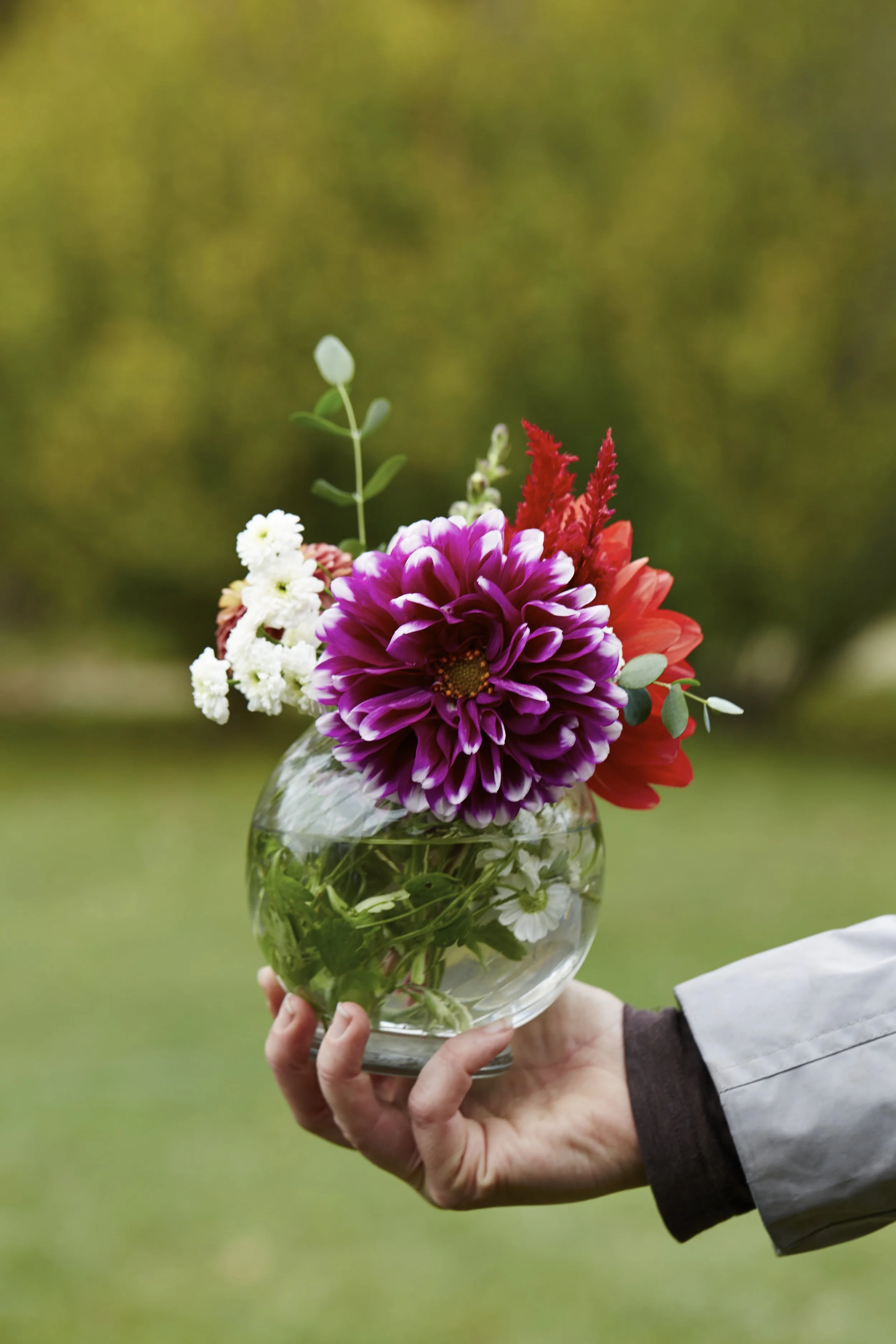 Person holding a glass bowl with colorful flowers including purple, white, pink, and red blooms, set outdoors with a blurred green background.