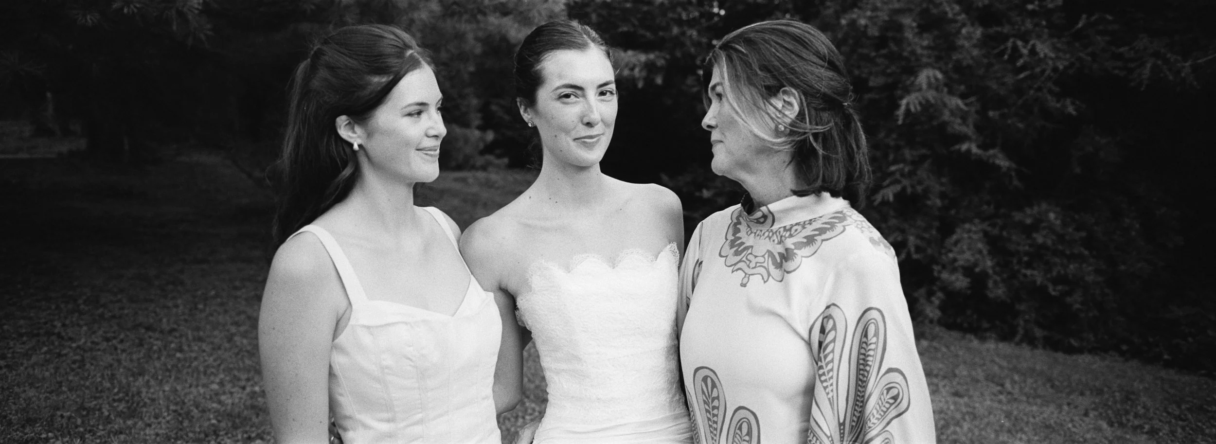 Three women standing outdoors, two of them dressed in wedding attire, facing each other and smiling, with trees in the background.