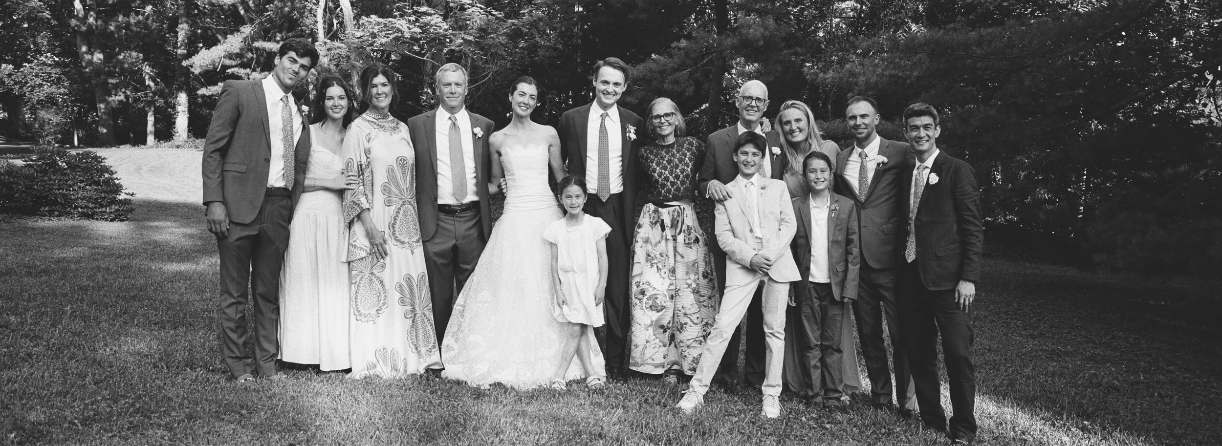 Group of people outdoors, dressed formally, standing on grass near trees, smiling for a photo.