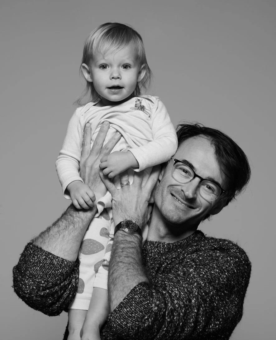 A man with glasses smiling while holding a young girl above his head in a studio setting, black and white image.