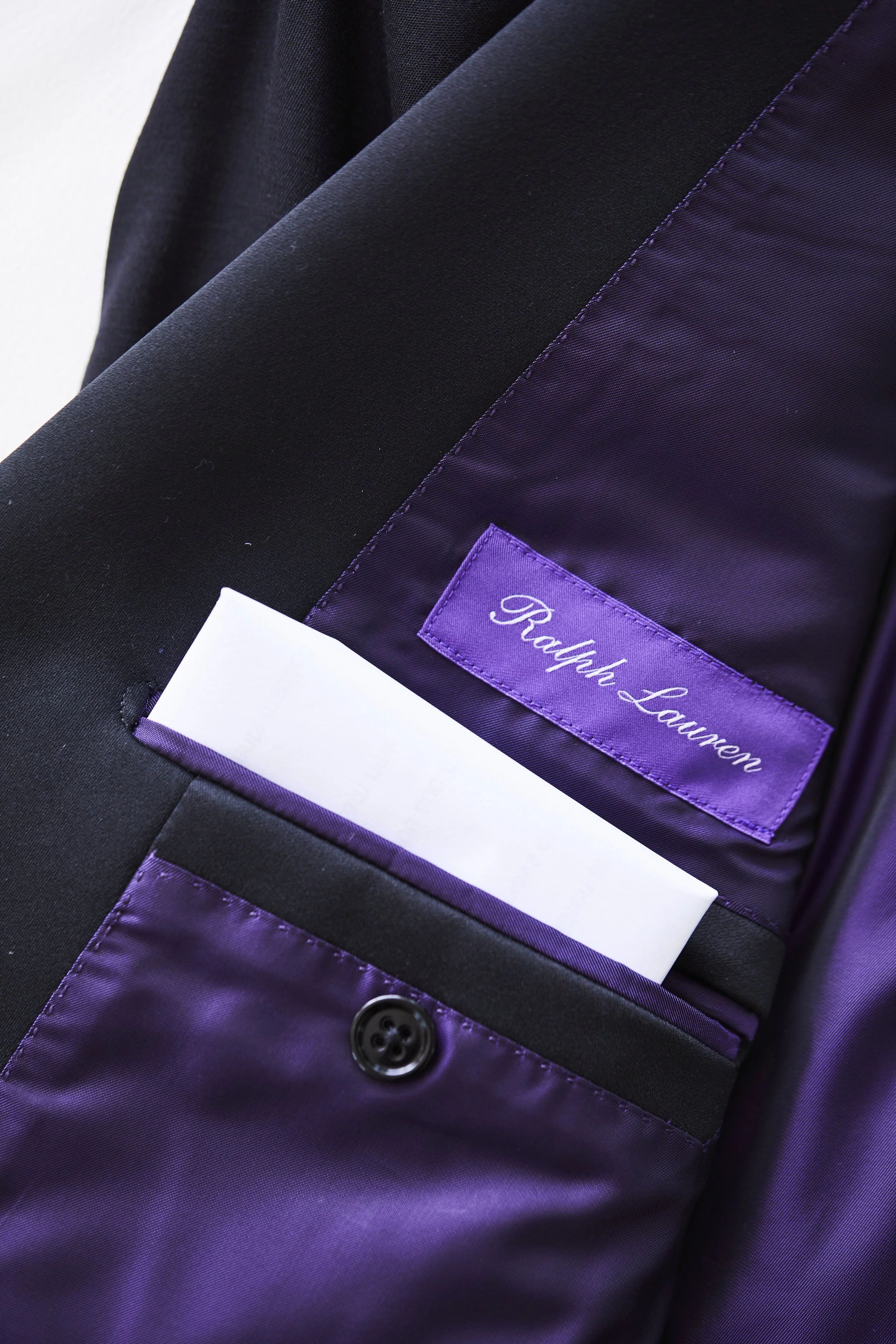 Close-up of the inside of a black suit jacket showing a purple lining, a white pocket square, a purple name tag with the embroidered name Ralph Lauren, and a black button.