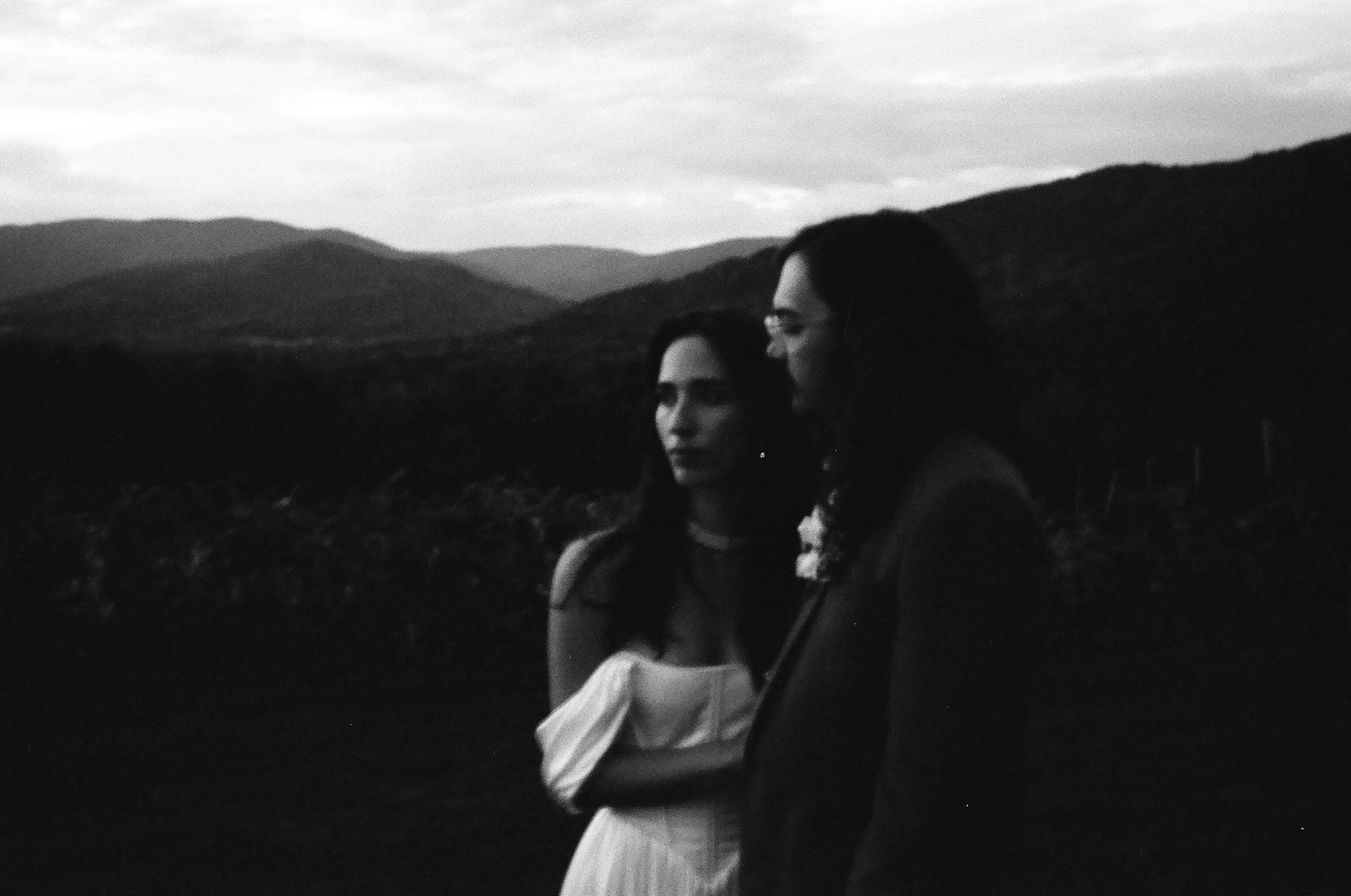Black and white photo of two women standing outdoors during dusk or dawn, with rolling hills and cloudy sky in the background. One woman is wearing a sleeveless dress, and the other is dressed in a blazer and glasses.