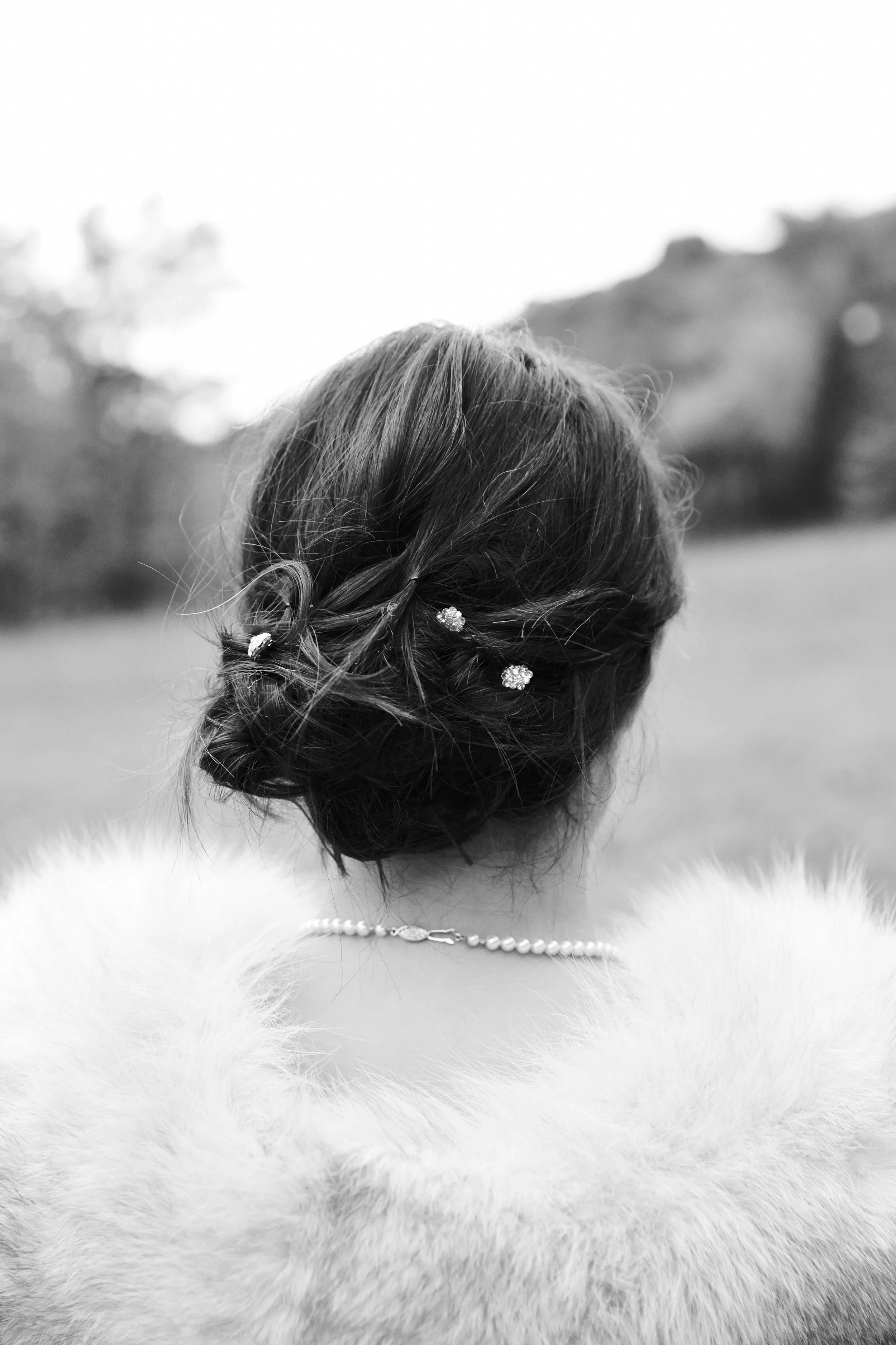 Black and white photo of a woman seen from behind, with her hair styled in an elegant updo adorned with small decorative hairpins, wearing a pearl necklace and a fur stole.