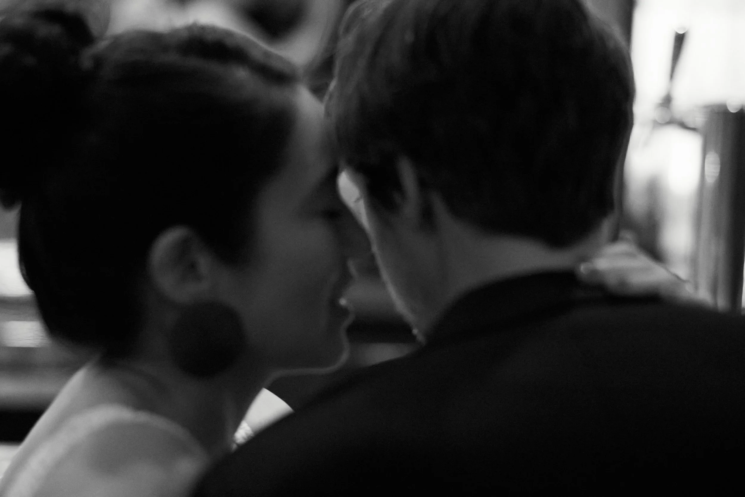 A black and white photo of a woman and a man in close proximity, appearing to be whispering or sharing an intimate moment.