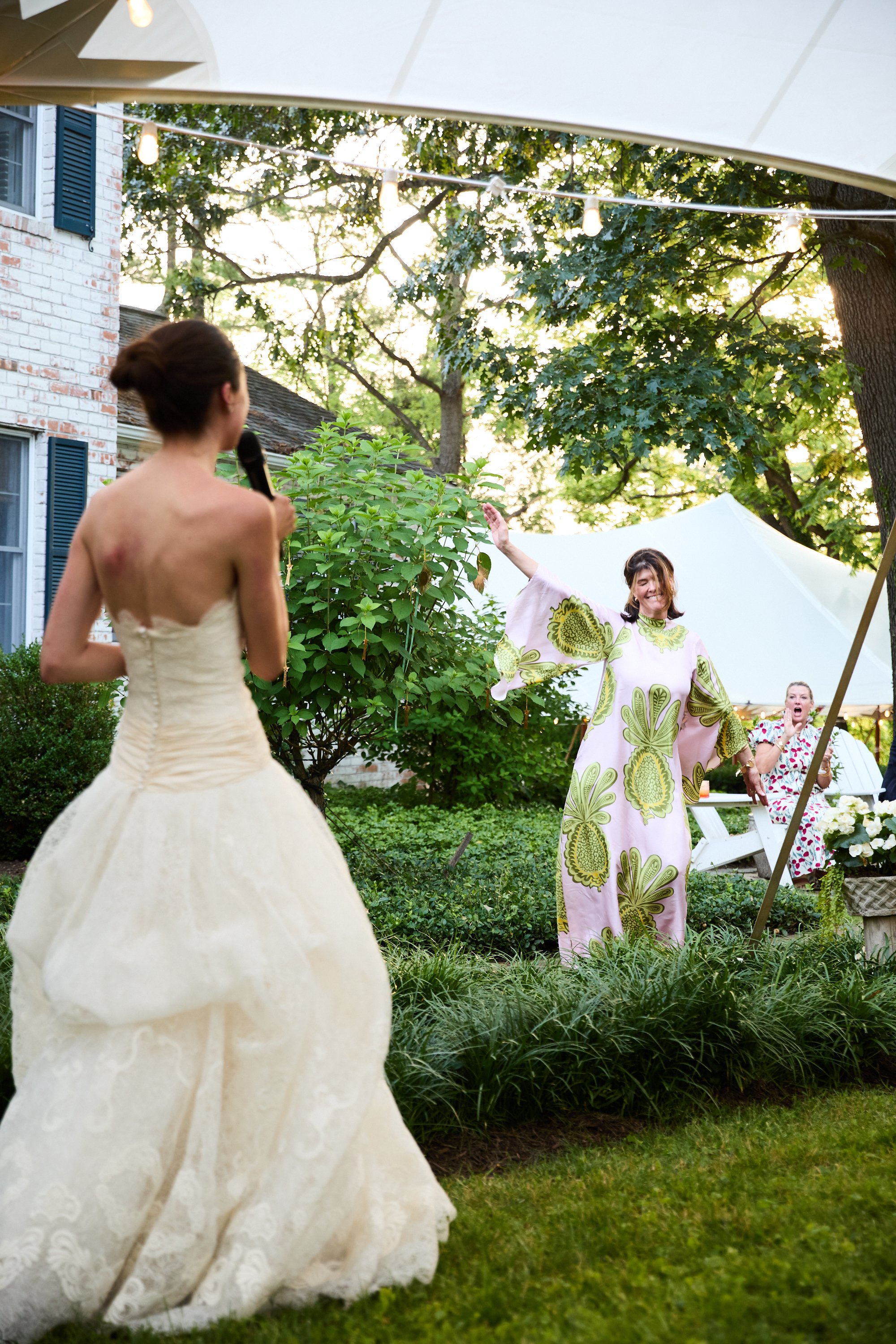 A bride in a strapless wedding gown standing in a garden, holding a microphone and speaking or singing to two women sitting under a white canopy, one of whom is wearing a colorful patterned dress and smiling, while the other looks surprised.