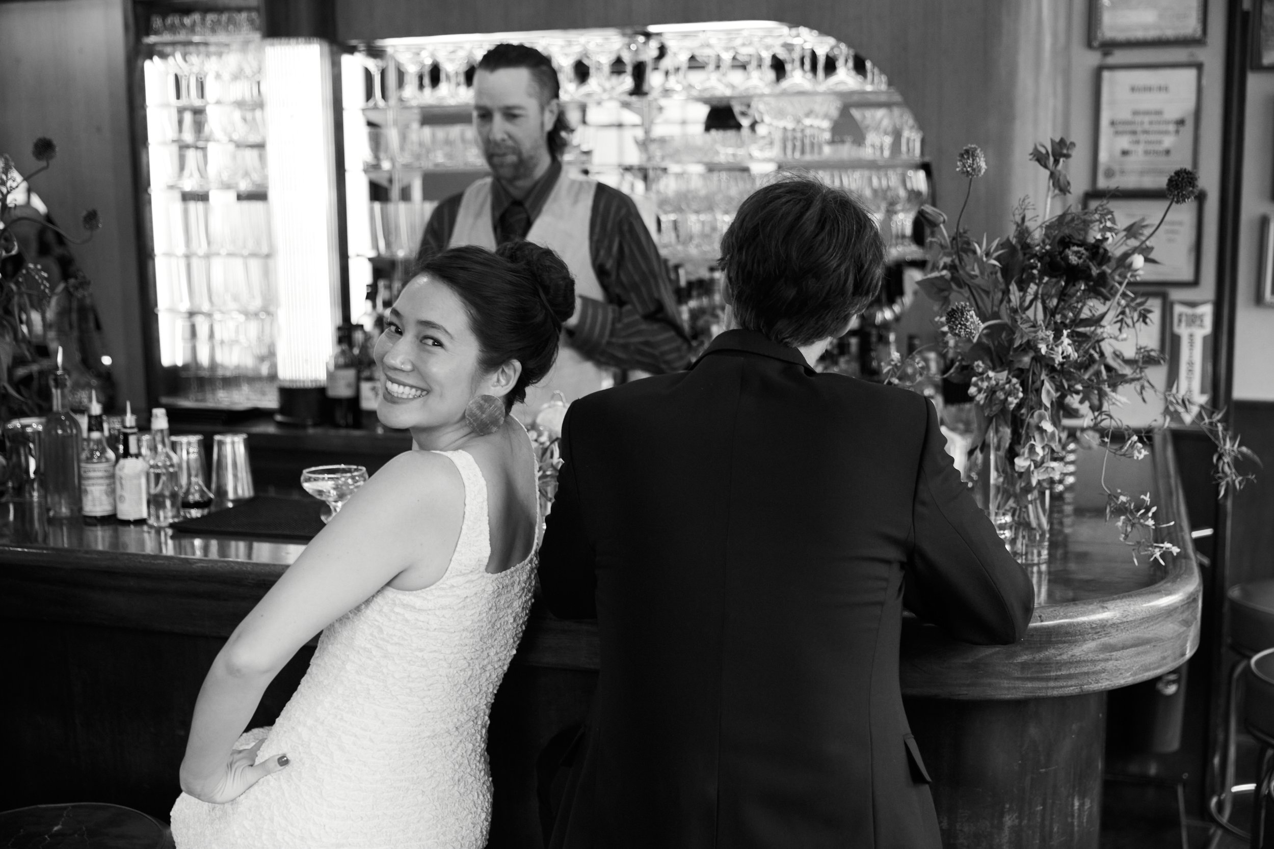 A black and white photo of a woman in a white dress smiling at the camera, seated at a bar with a man in a suit sitting beside her with his back to the camera. A bartender is behind the bar, and there is a large flower arrangement on the counter.