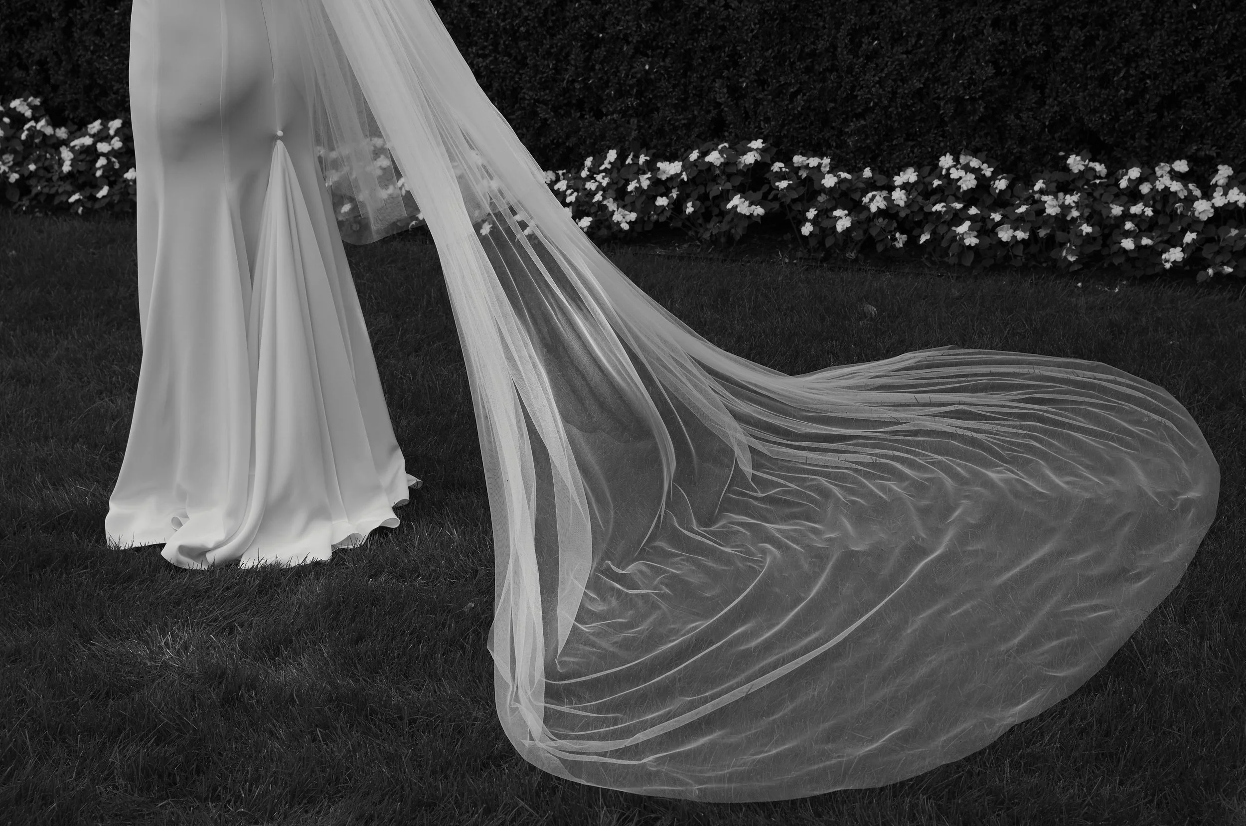 A bride in a wedding dress with a long veil standing on grass near a flower bed.