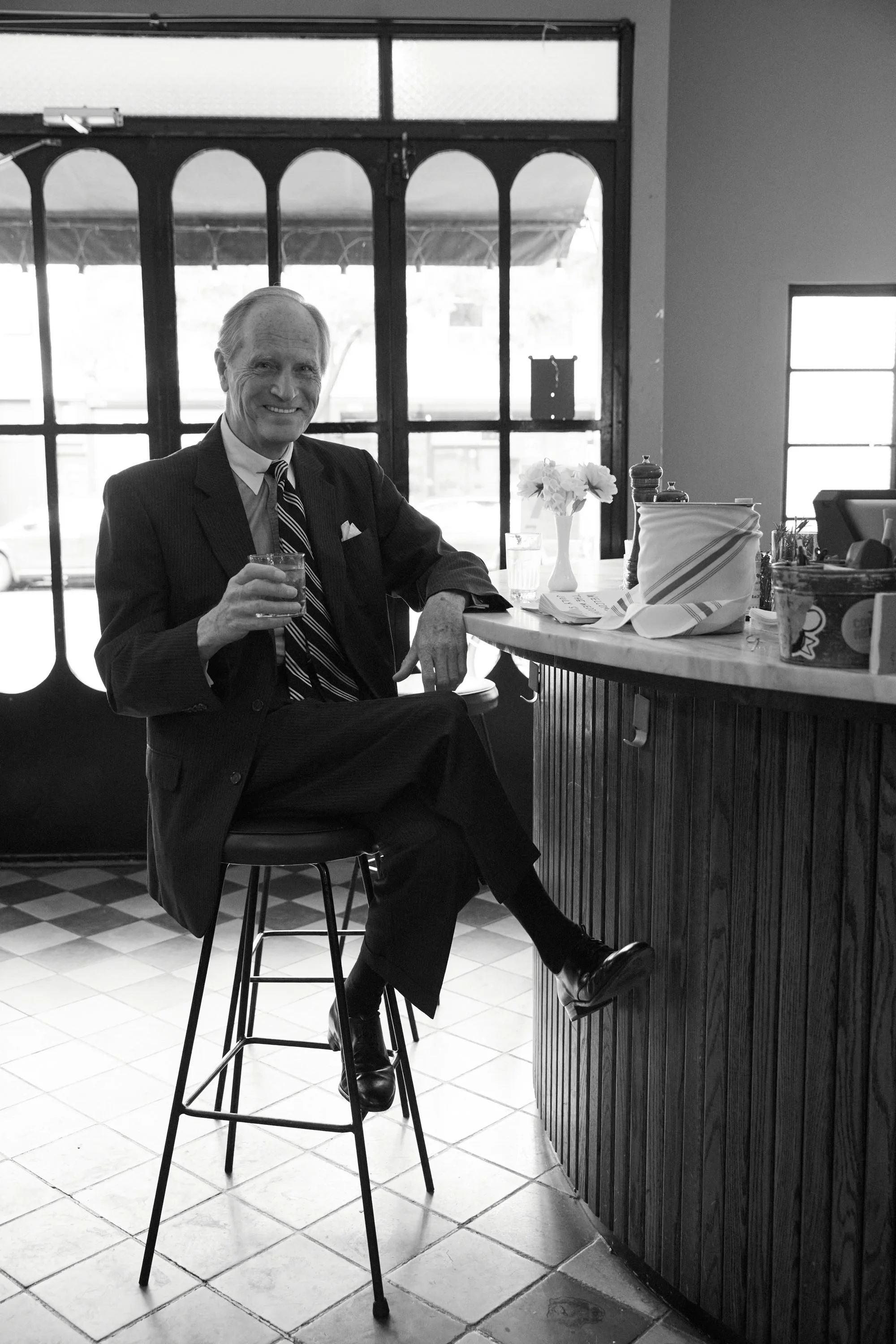 A man in a suit sitting on a barstool at a counter, holding a drink, smiling, in a cafe or restaurant with large windows in the background.