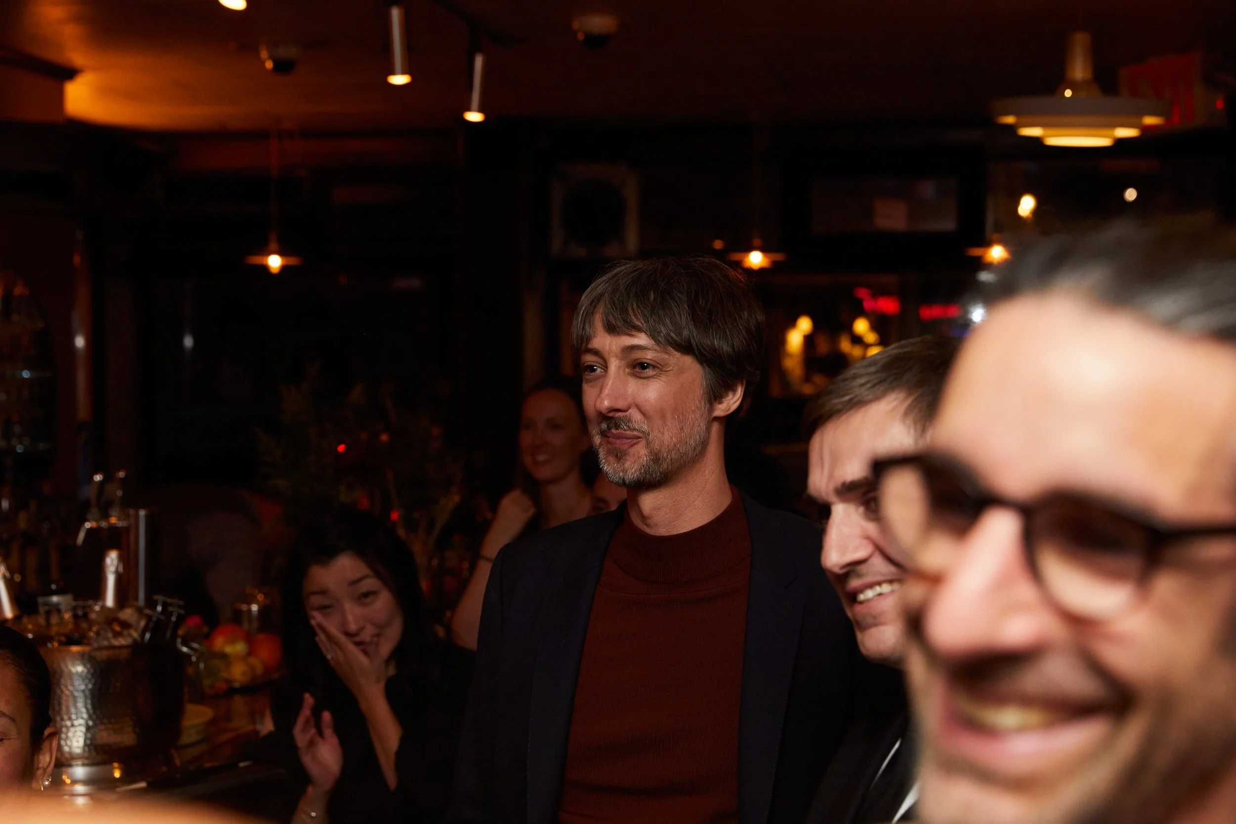 A group of people at an indoor gathering, some smiling and laughing, in a dimly lit setting with warm lighting.