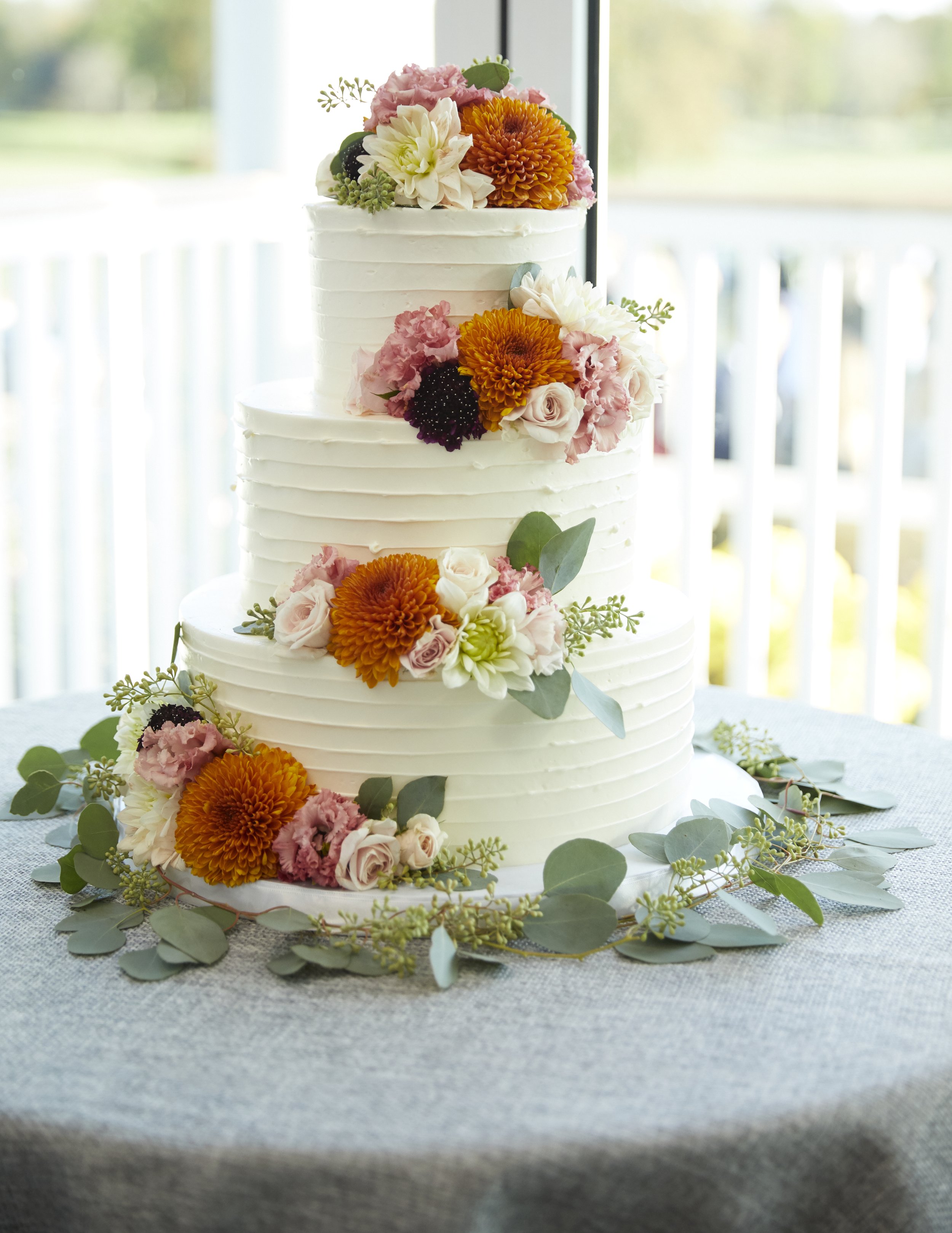 Three-tier white wedding cake decorated with orange, pink, and white flowers, surrounded by greenery, on a gray tablecloth.