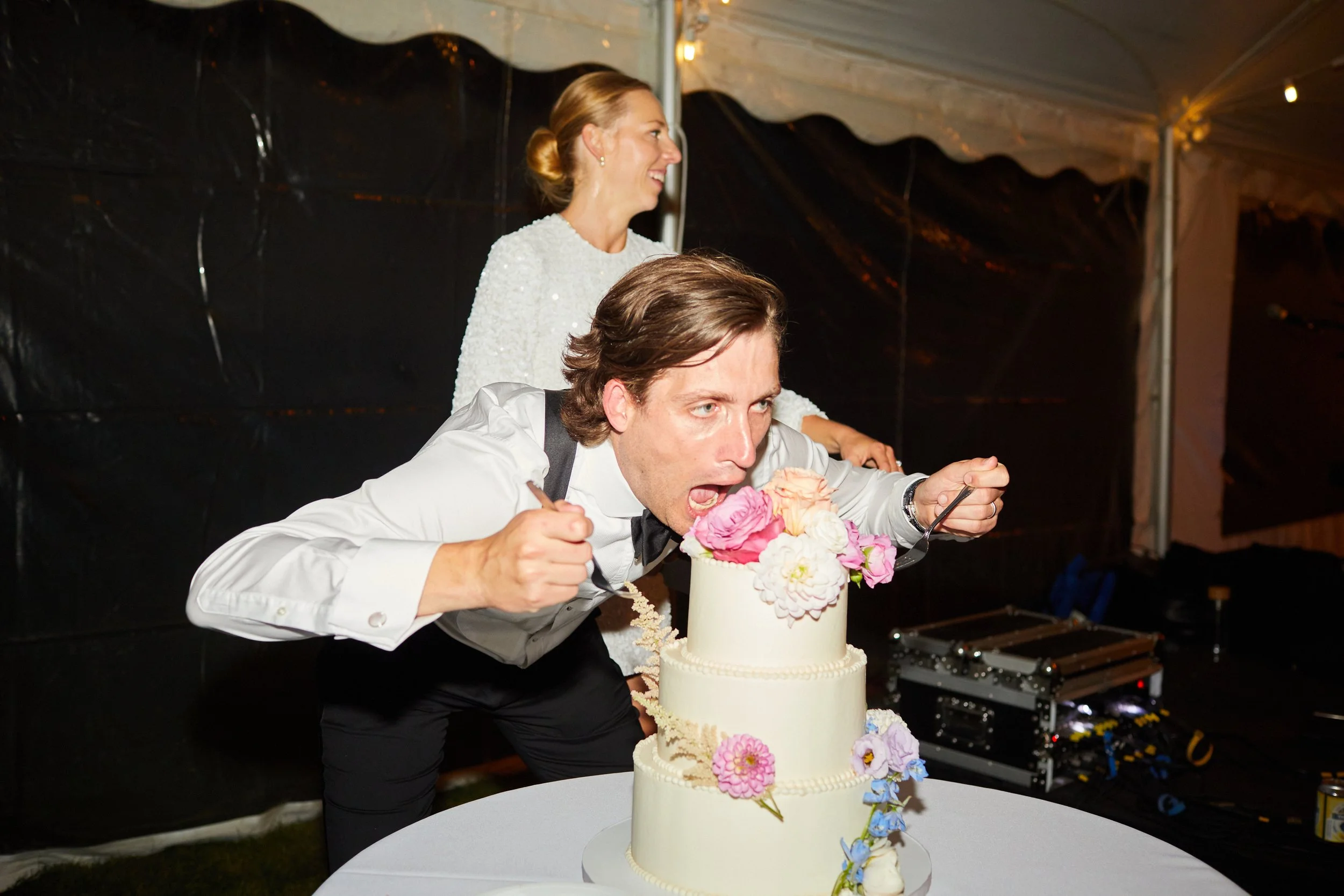 A man in a tuxedo and a woman in a white dress pretend to eat a multi-tiered wedding cake decorated with pink, white, and purple flowers at a wedding reception.