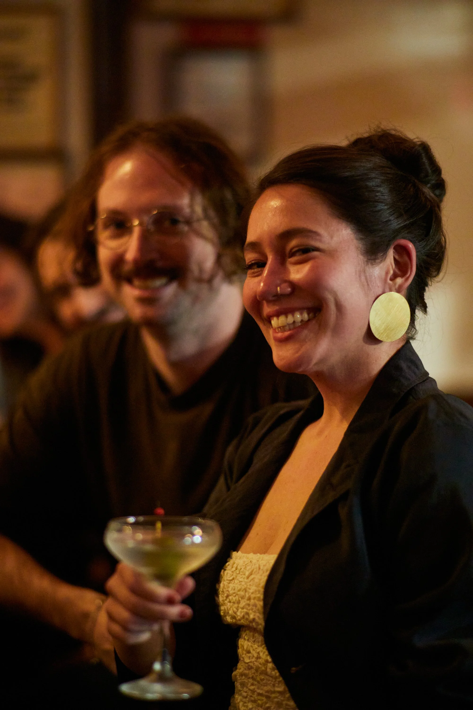 Two people, a woman and a man, at a social gathering. The woman is holding a cocktail glass and smiling, wearing large round earrings, with her hair styled up. The man, with glasses and shoulder-length hair, is smiling in the background.