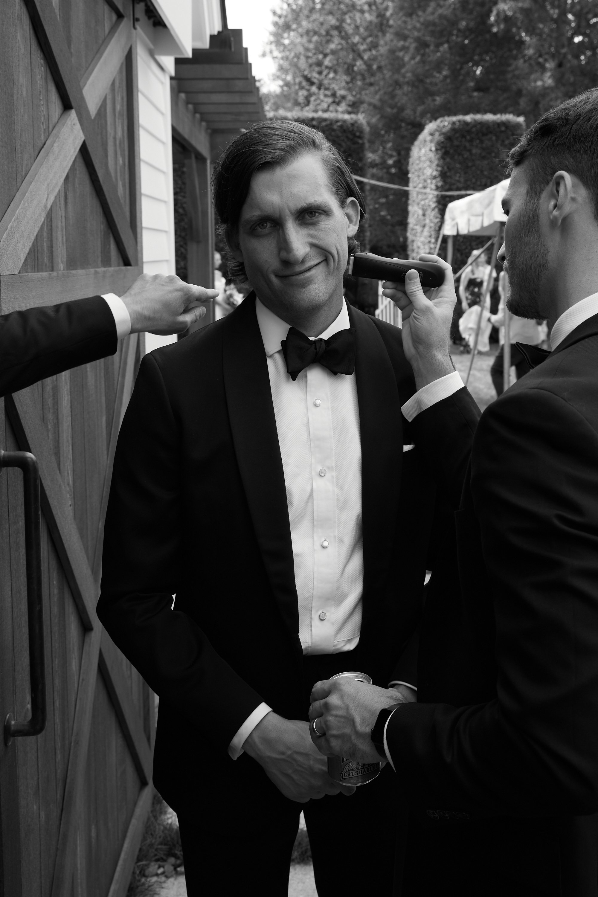 Black and white photo of a man in a tuxedo getting a beard trimmed with an electric razor at an outdoor event.