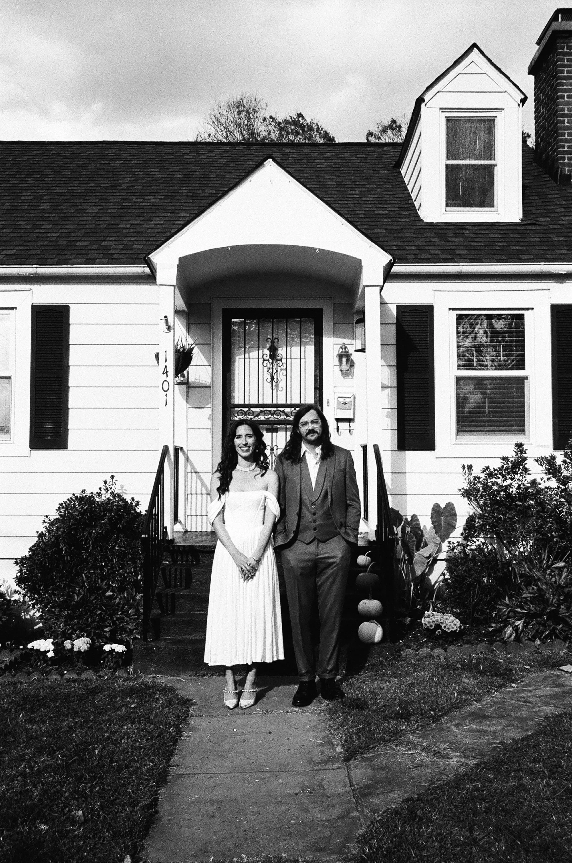 A couple standing in front of a house, dressed in vintage attire, smiling, with plants and pumpkins nearby in black and white.