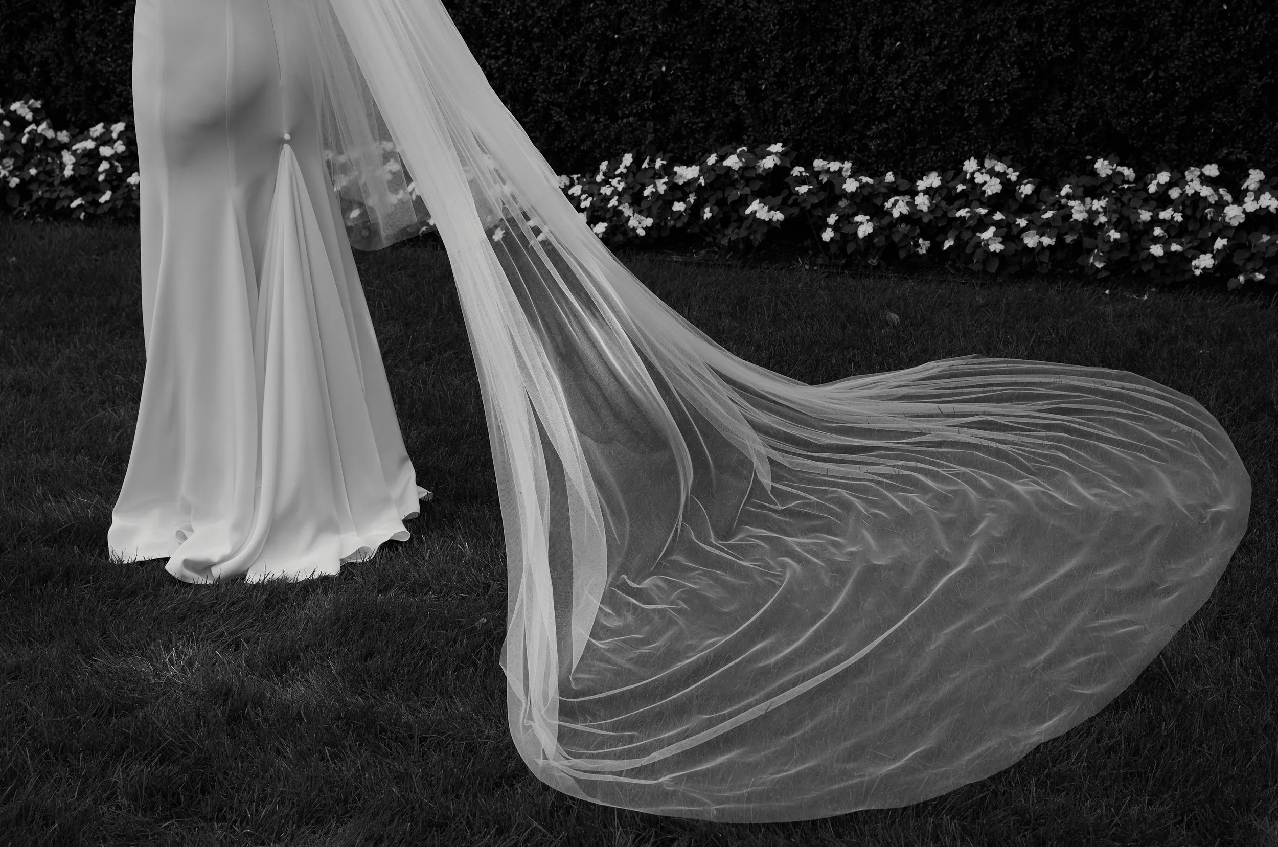 Close-up of a wedding dress with a long train and veil on a grassy area with flower bed in background.