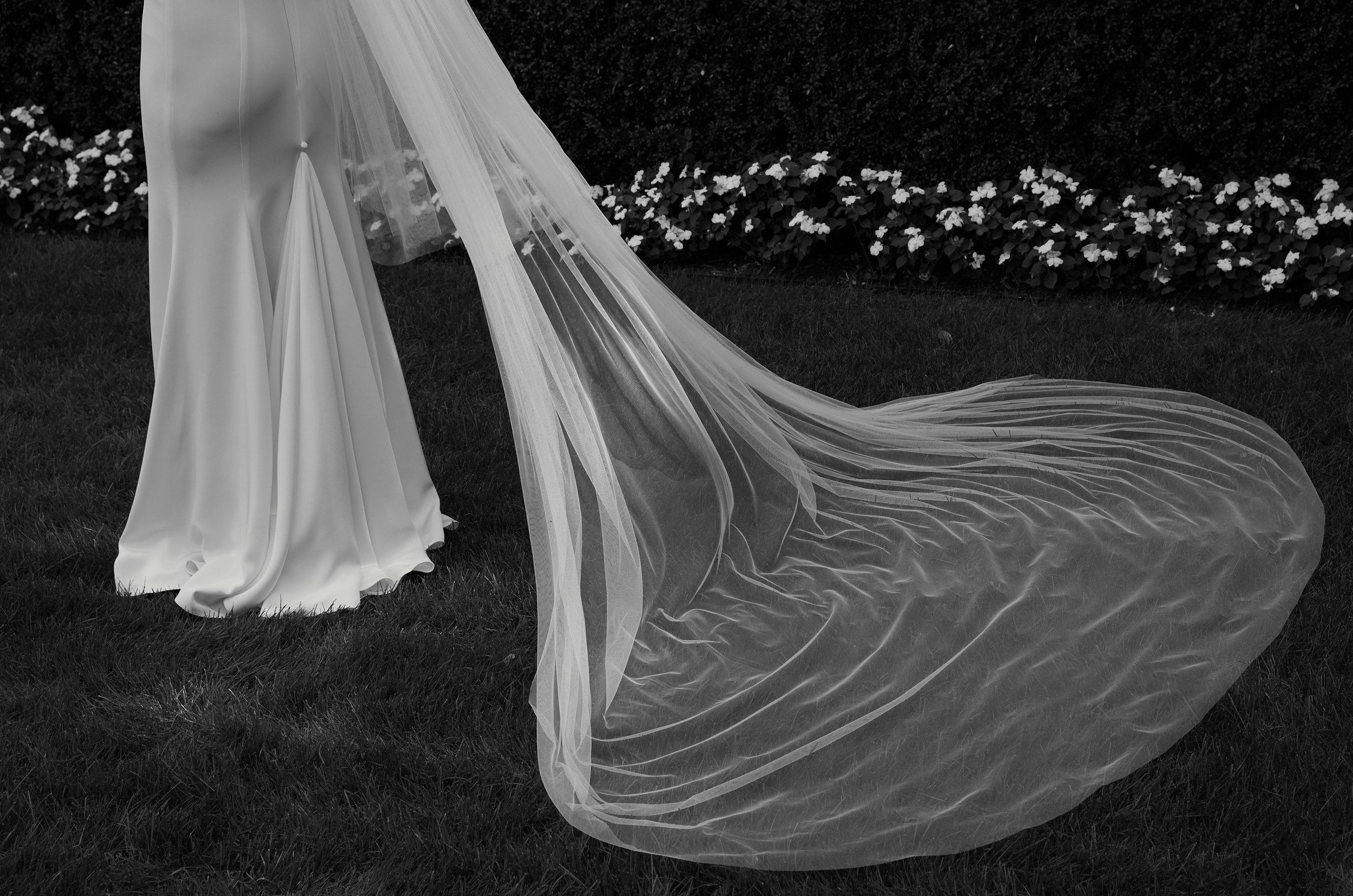 Close-up of a wedding dress and train on grass, with hedge and flowers in background.