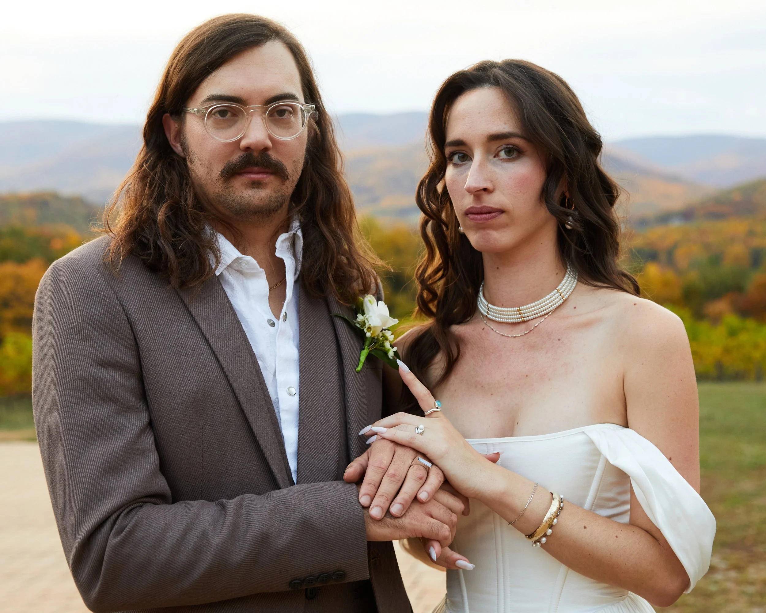 A couple dressed in wedding attire standing outdoors with mountains and autumn foliage in the background.