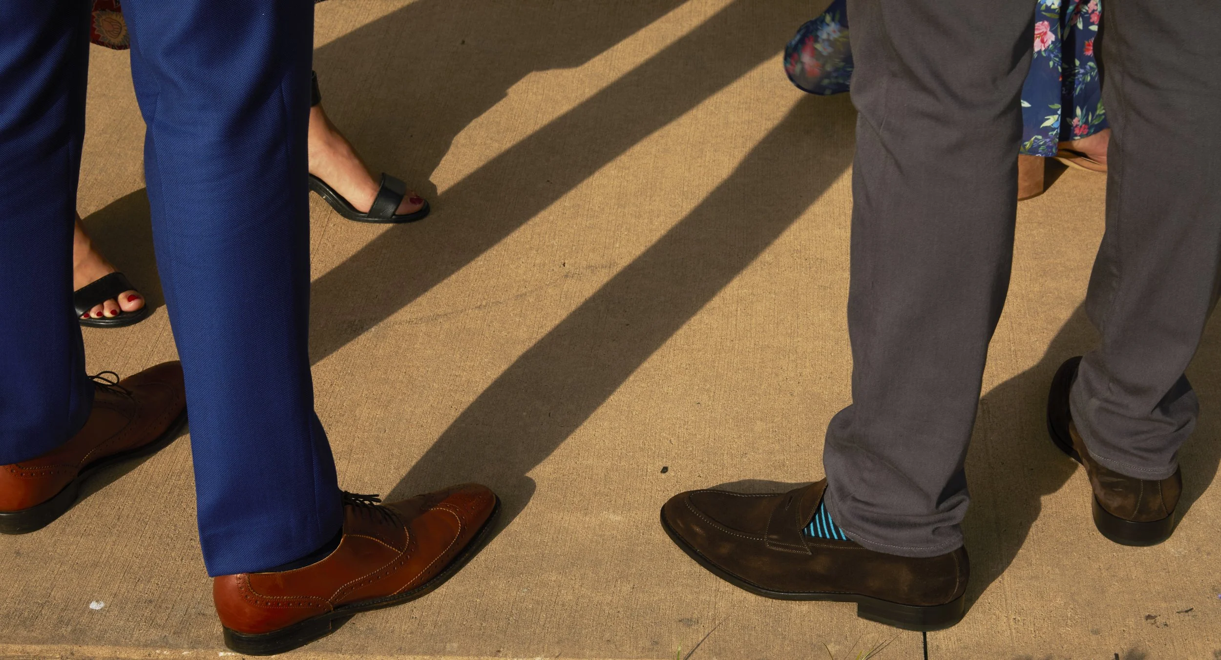 Close-up of the lower half of two people standing on a beige carpet, showing their legs and footwear. One person is wearing navy blue pants and brown dress shoes, while the other is wearing gray pants and dark brown shoes with blue striped socks. Sha