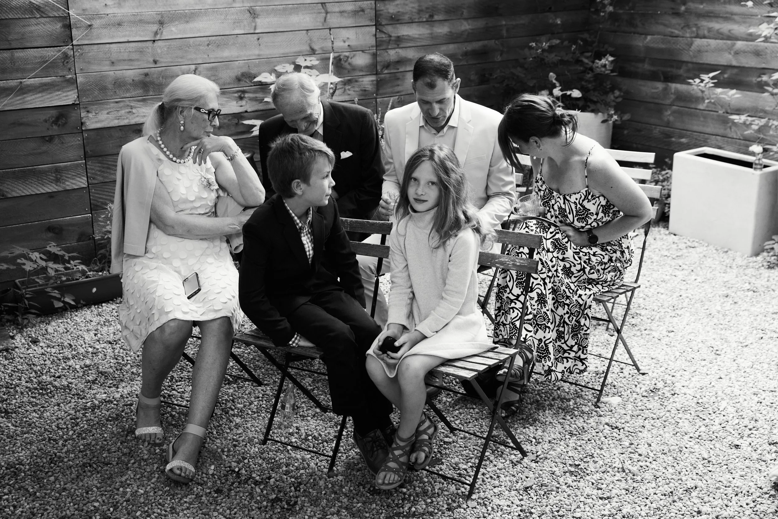 Group of six people sitting outdoors on chairs near a wooden fence, engaged in conversation, in black and white.
