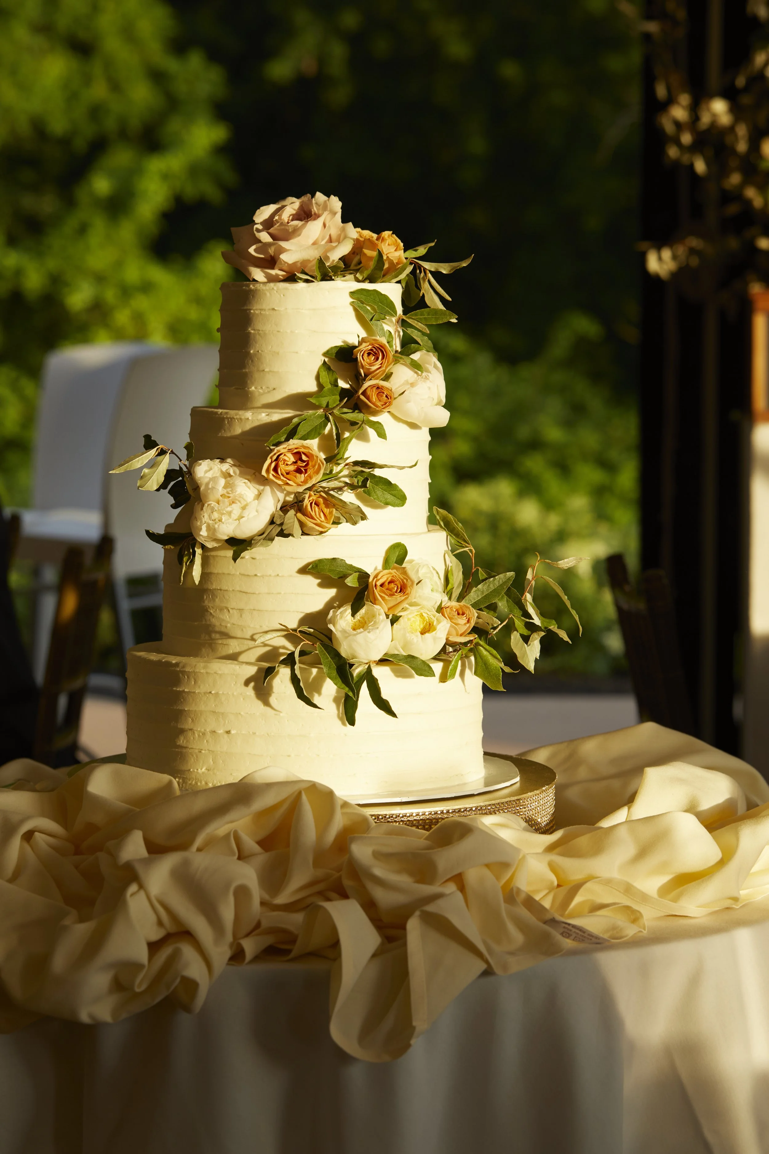 A four-tiered wedding cake decorated with fresh flowers including roses and peonies, set on a table with cream-colored fabric, outdoors with greenery in the background.