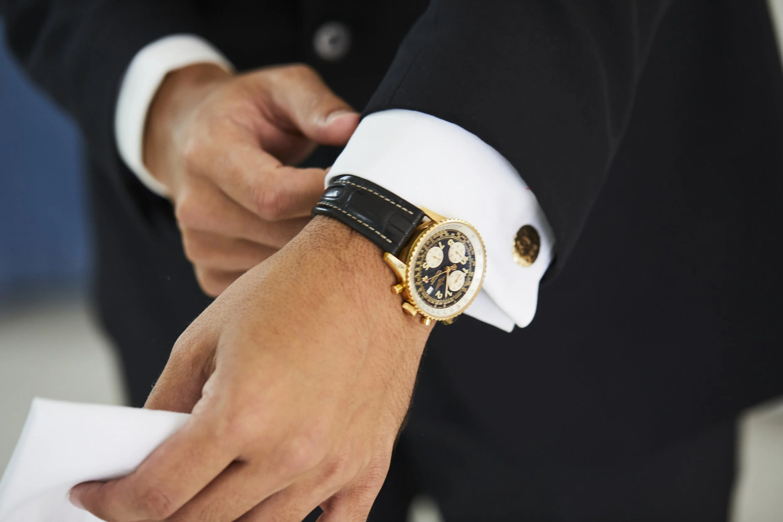 Close-up of a man's wrist with a gold watch, wearing a dark suit jacket and white shirt with cufflinks, adjusting his cuff.