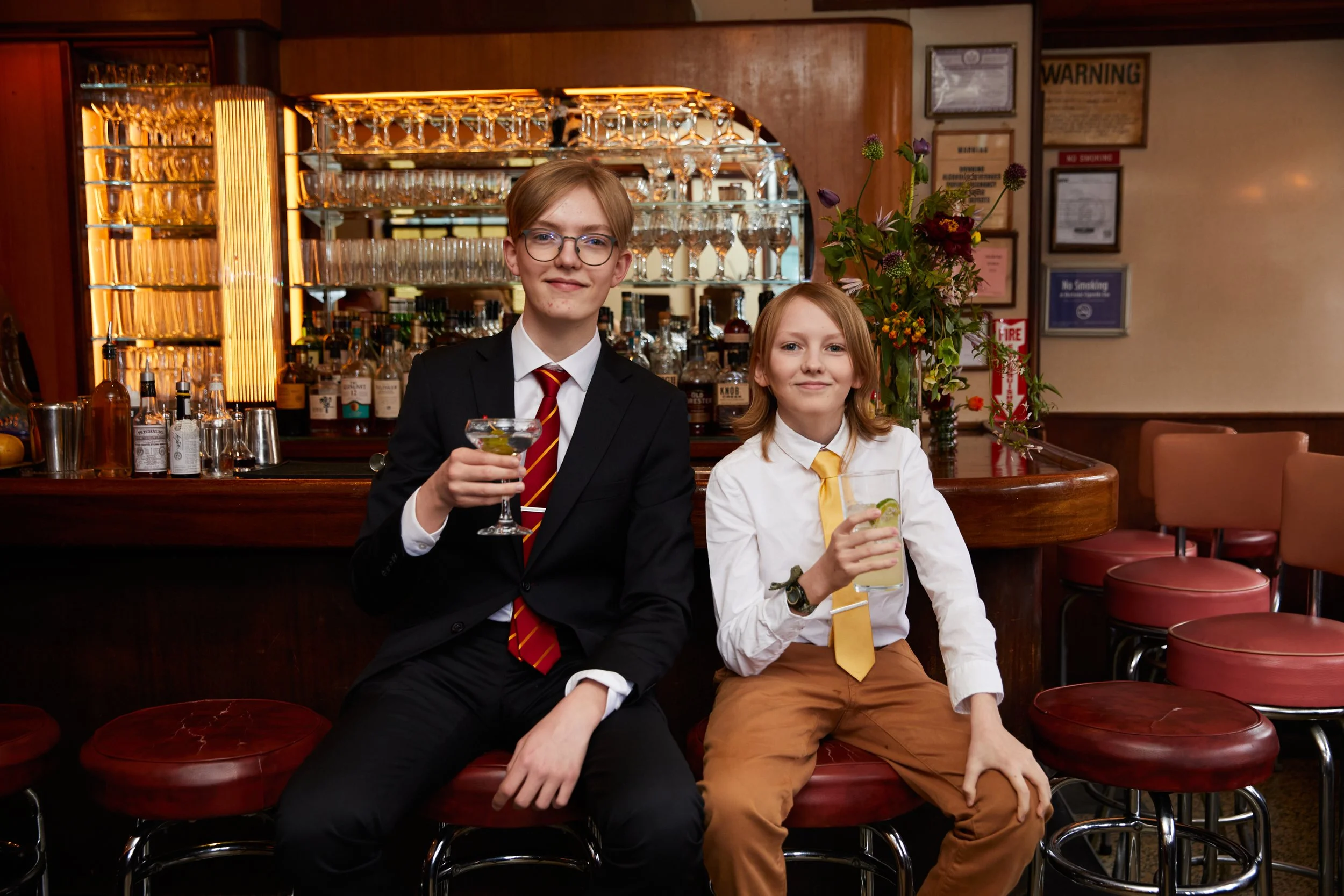 Two young boys dressed in formal attire sitting at a bar counter, each holding a drink, in a dimly lit bar or restaurant setting with shelves of liquor bottles and glassware in the background, and a large flower arrangement nearby.