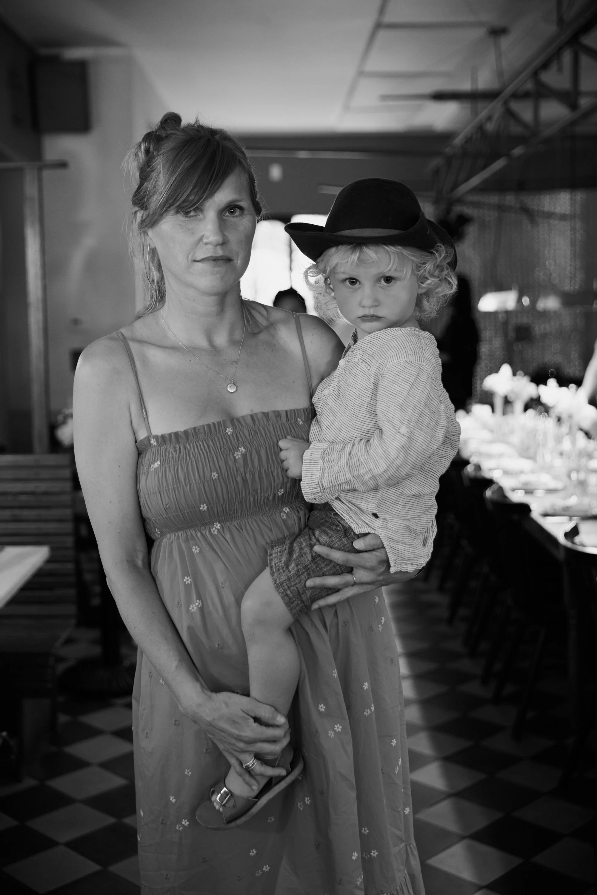 A woman holding a young child with curly hair and a cowboy hat in a restaurant or event space with tables and chairs.