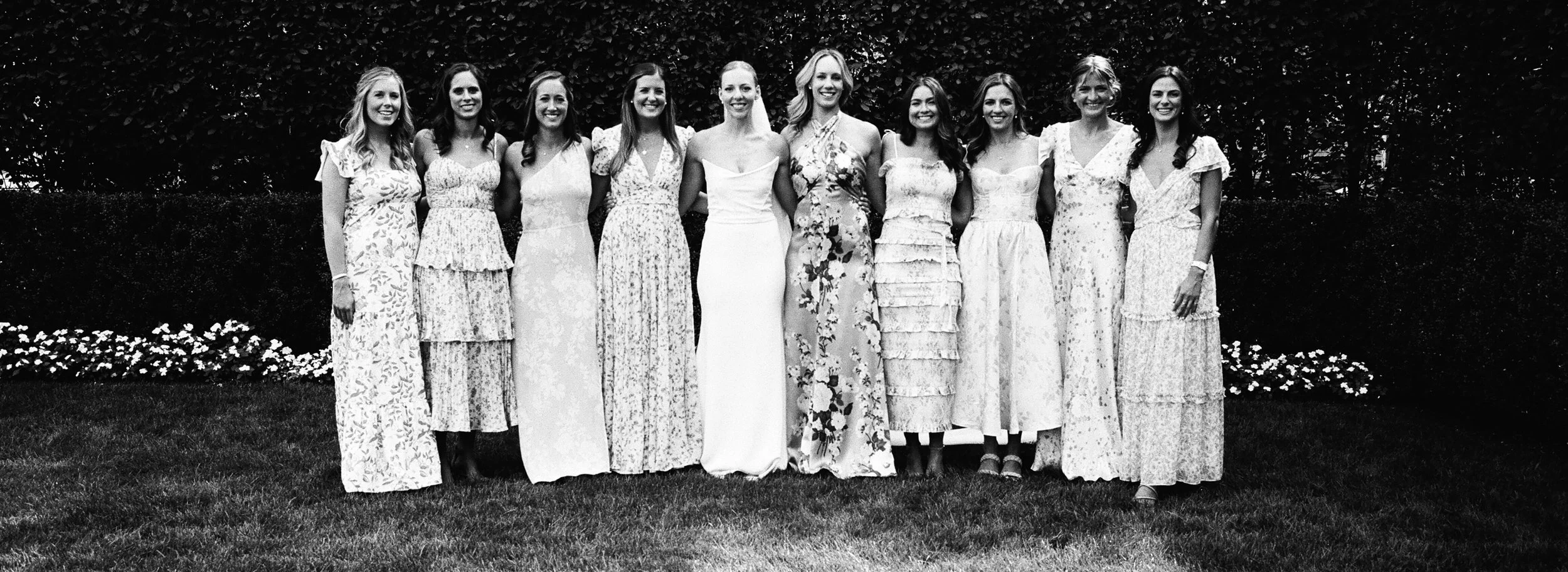 A group of eleven women standing outdoors on grass in front of bushes, dressed in long floral or solid dresses, smiling for a photo.