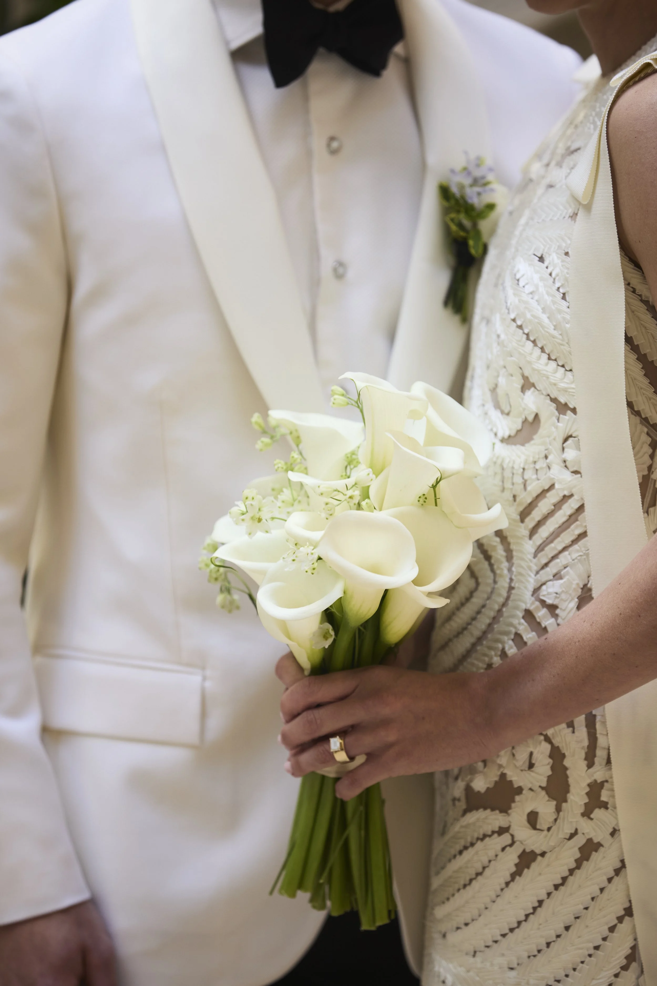 Close-up of a bride holding a bouquet of white calla lilies and small white flowers, standing beside a groom dressed in a cream tuxedo with a black bow tie.