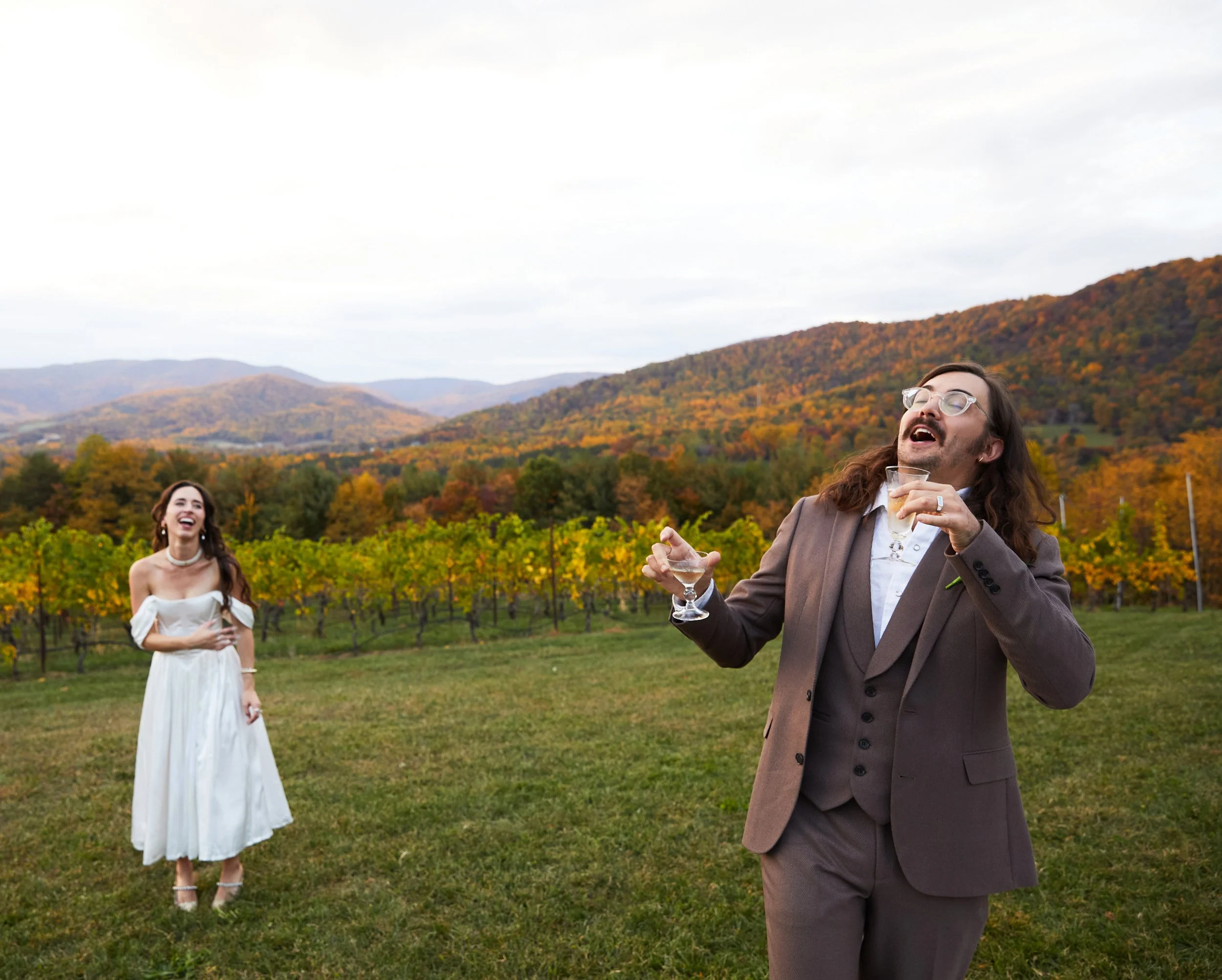 A man in a brown suit and glasses holding a drink and laughing, standing in front of a vineyard with a woman in a white dress, smiling and standing with her hand on her stomach, with colorful autumn mountains in the background.