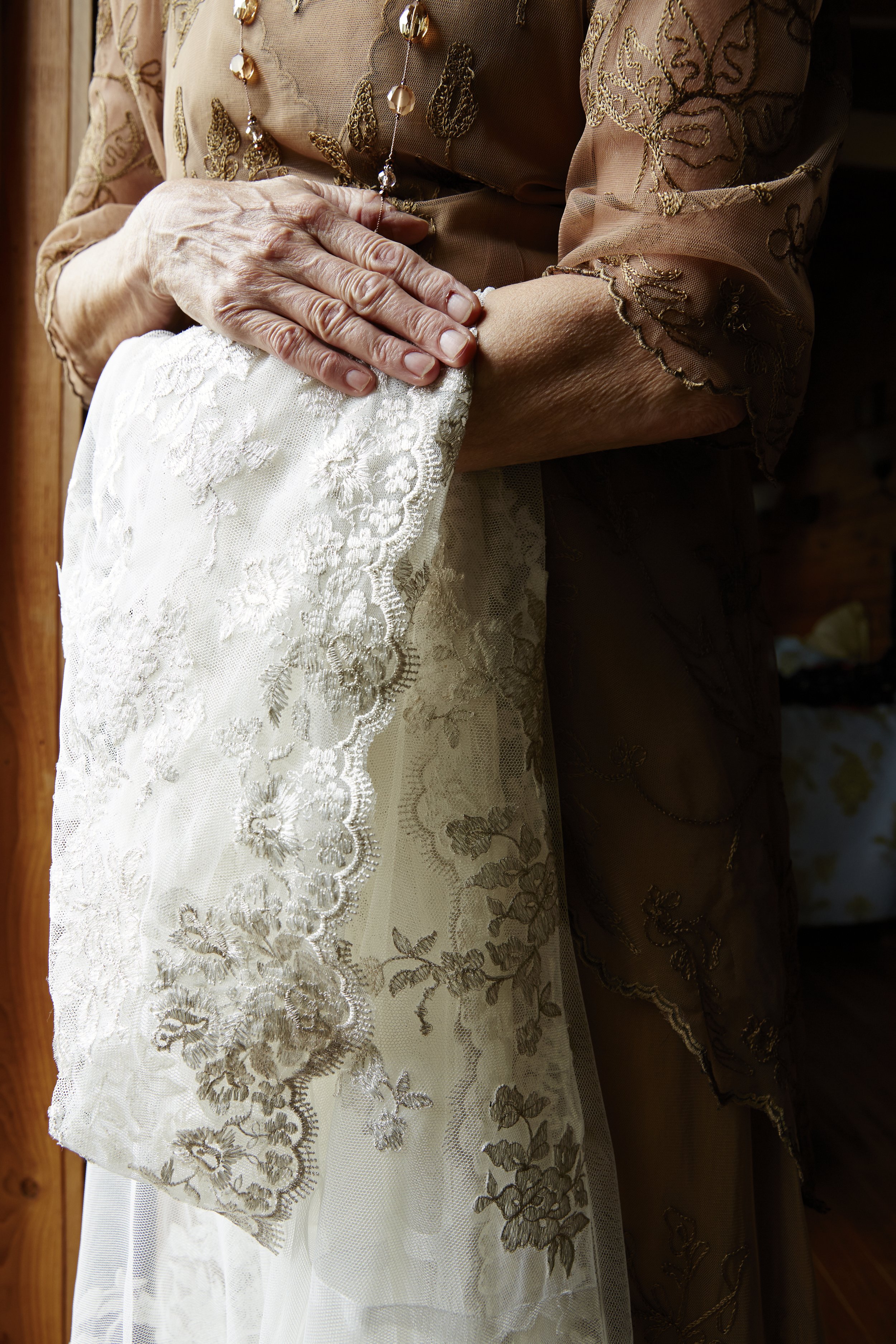 Close-up of elderly woman's hands resting on a white lace curtain or fabric, wearing a light brown, embroidered blouse with intricate gold detailing.