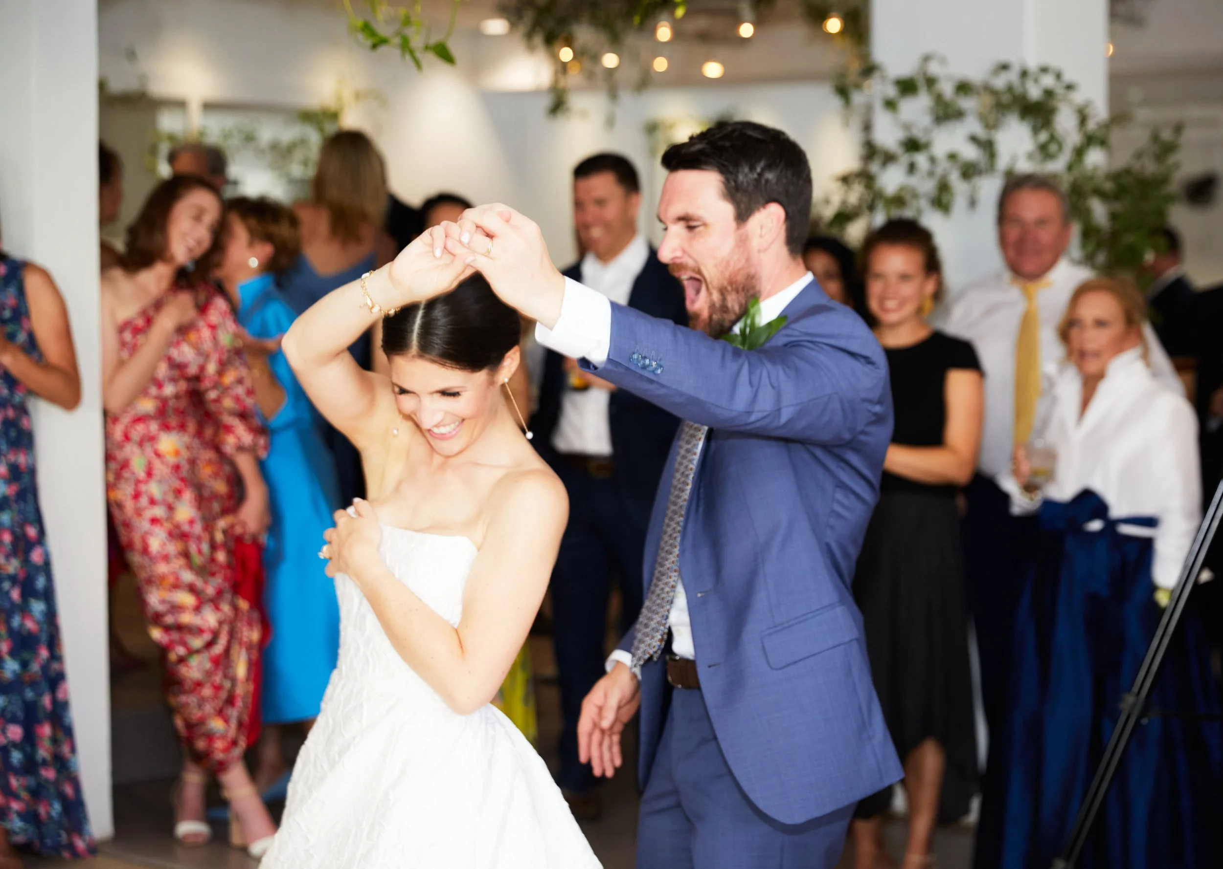 A newlywed couple is dancing at their wedding reception, surrounded by friends and family. The bride is smiling and wearing a white strapless wedding dress, and the groom, in a blue suit, is passionately dancing with her. Guests are watching and enjo