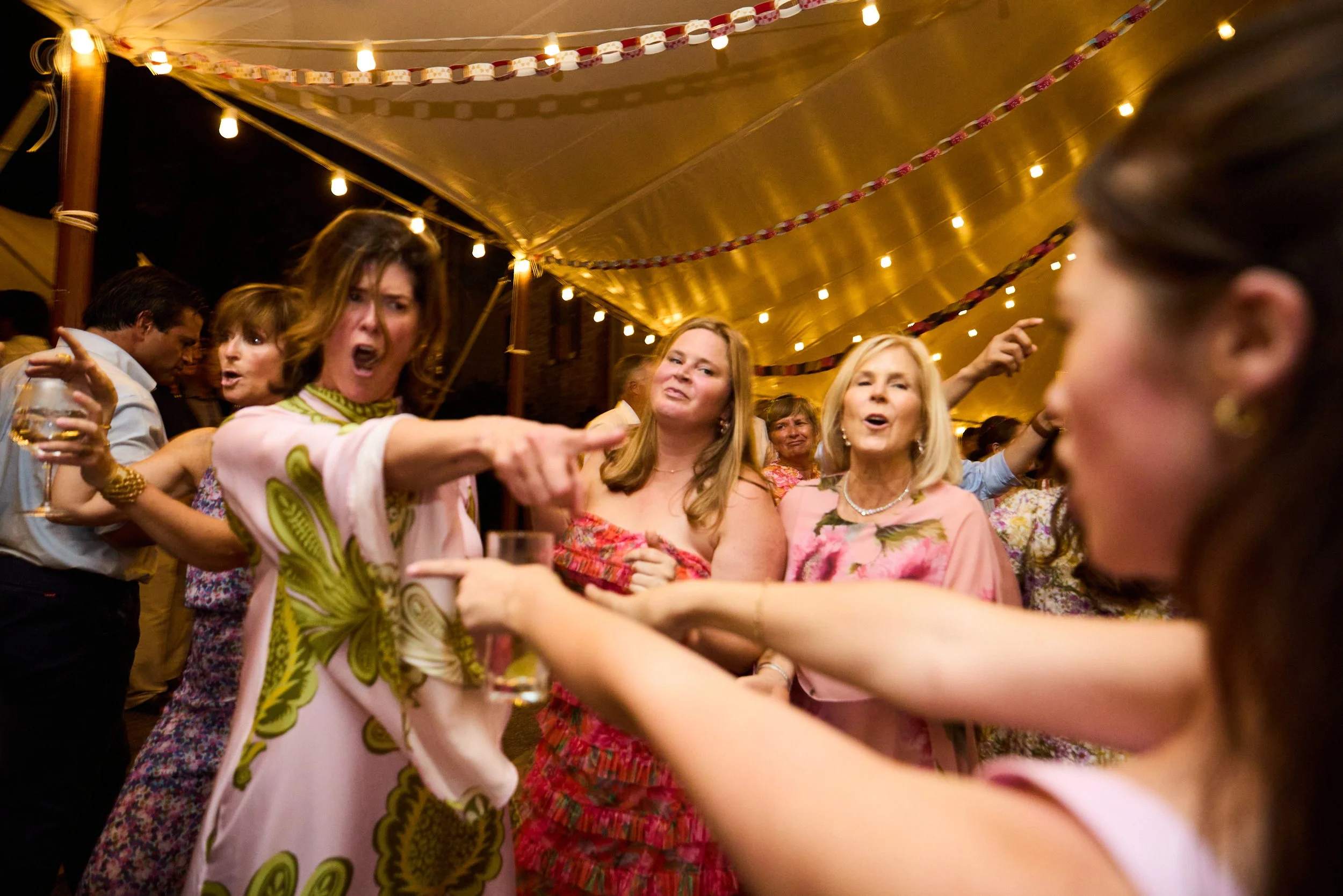 Group of women at a lively party under yellow canopy with string lights, engaged in conversation and holding drinks.