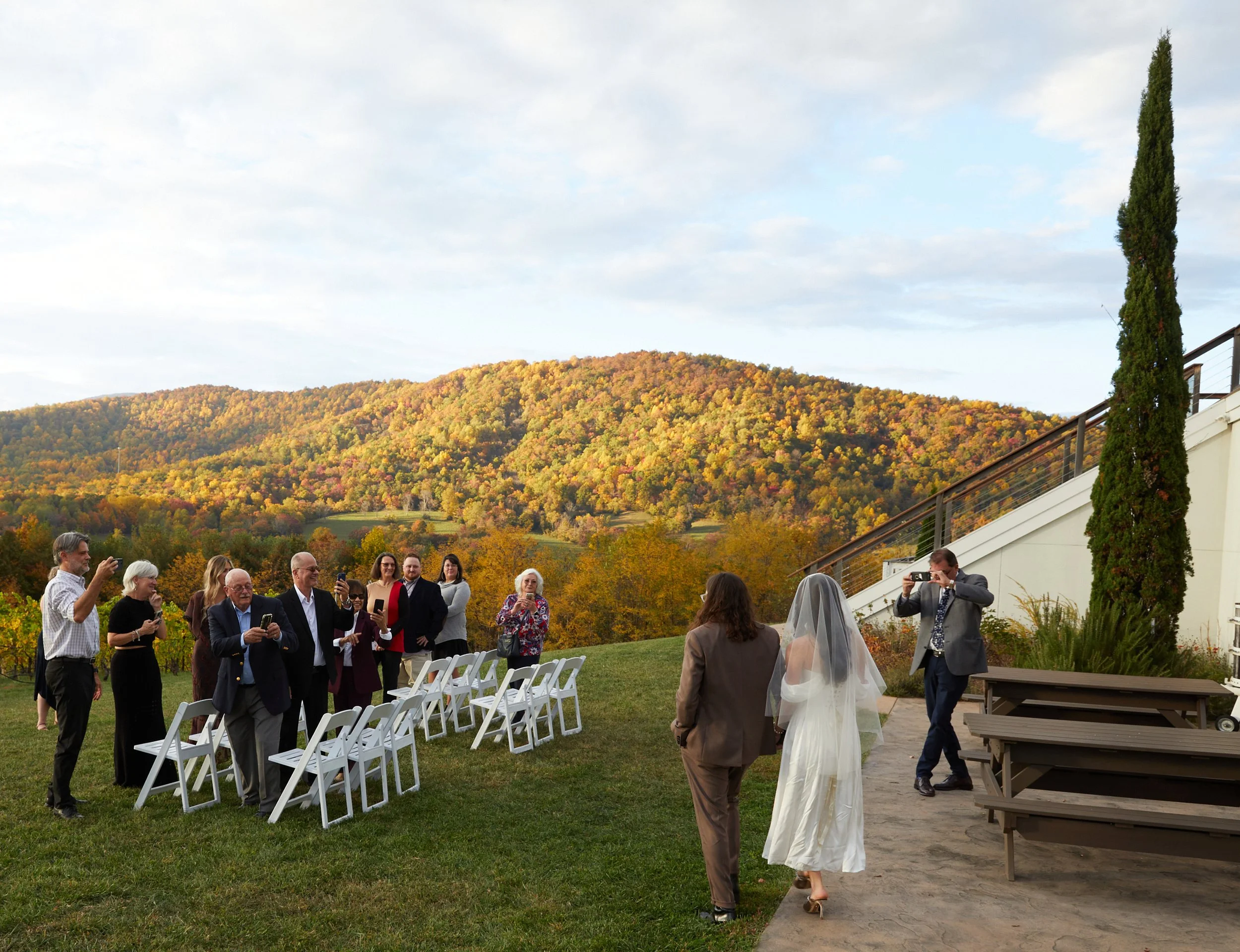 A wedding ceremony outdoors with guests standing and taking photos, a bride and guest walking towards the ceremony, set against an autumn landscape with hills and trees.